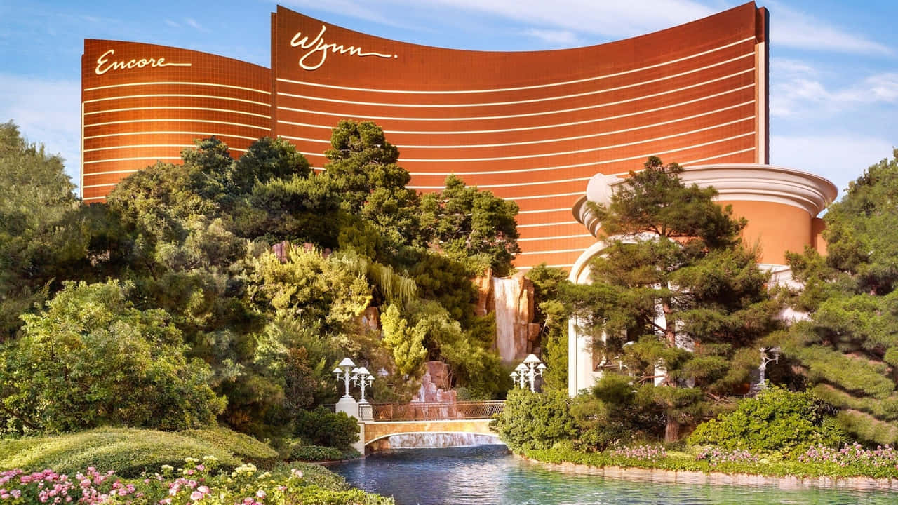 720p Las Vegas 桌布 壁紙 — 一座巨大的橙色弧形酒店，帶有「Wynn」和「Encore」標牌，聳立在茂密的綠色植物和寧靜池塘旁的層疊瀑布之上。
