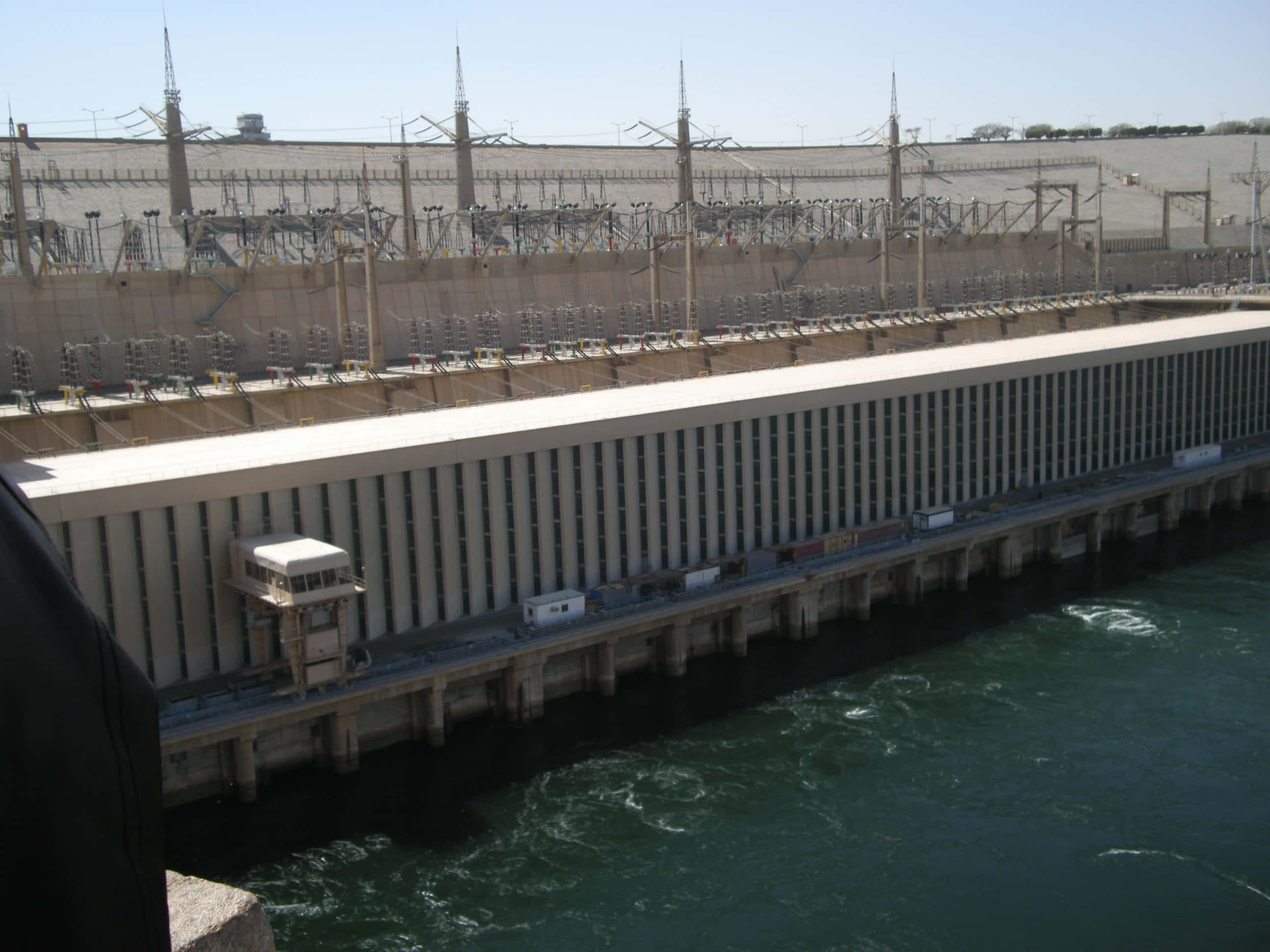 水 桌布 壁紙 — 一座巨大的混凝土水力發電大壩，帶有電線和渦輪機，在明亮的日光下釋放出湍急的綠色水流。