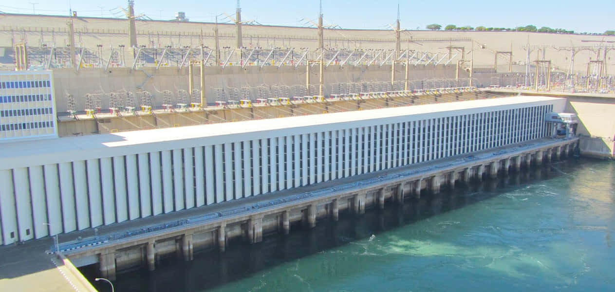 水 桌布 壁紙 — 一座巨大的混凝土水力發電大壩，設有渦輪機和輸電線路，在清澈的藍天下。