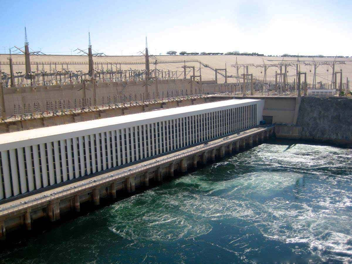 沙漠 桌布 壁紙 — 一座巨大的混凝土水力發電大壩在明亮的日光下釋放湍急的綠色水流，上方有工業輸電線。