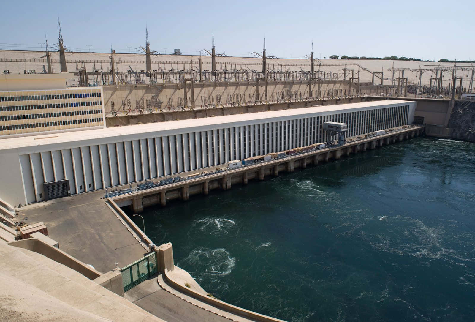 水 桌布 壁紙 — 一座巨大的混凝土水力發電水壩在明亮的日光下釋放湍急的深藍色水流，背景有工業電線和建築物。