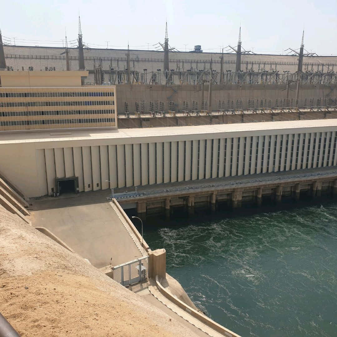 水 桌布 壁紙 — 一座巨大的混凝土水壩，設有發電廠建築和電塔，矗立在奔騰的綠色河流之上，天空晴朗。