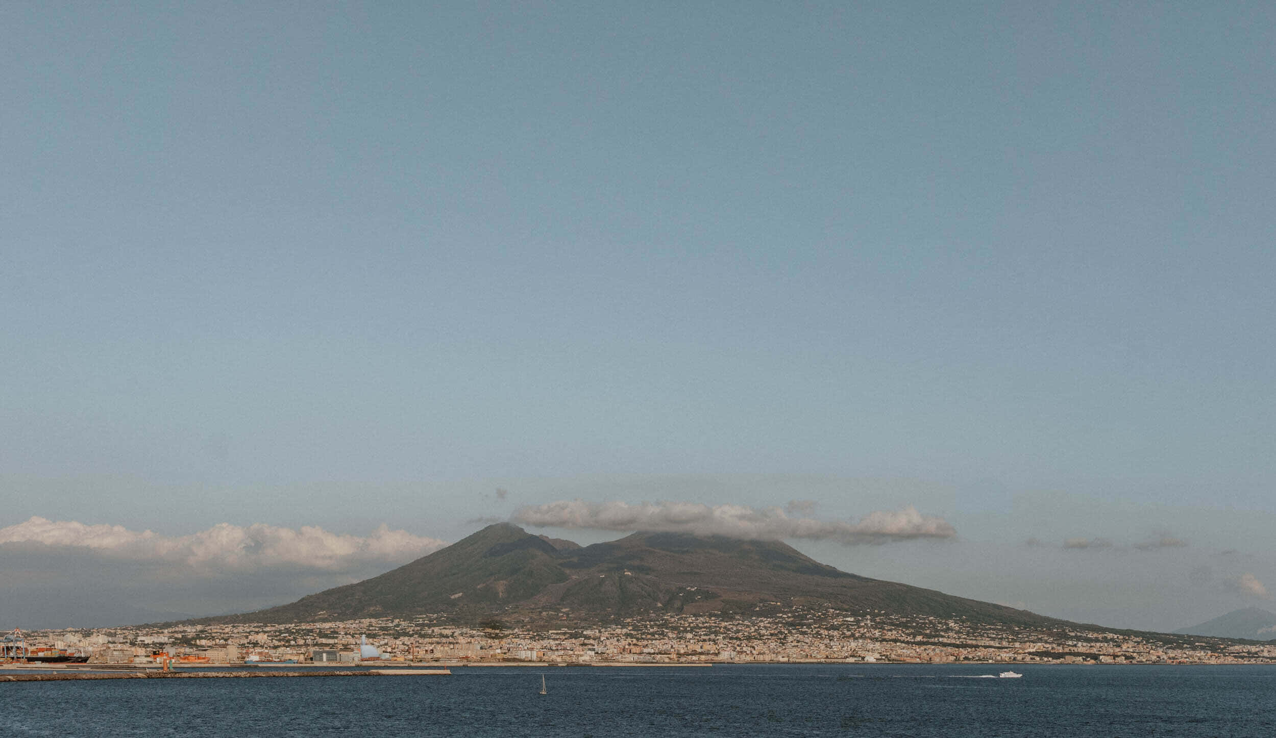 Volcano 桌布 壁紙 — 一座巨大的火山聳立在海岸城市景觀之上，天空淡藍，雲朵零星分佈。