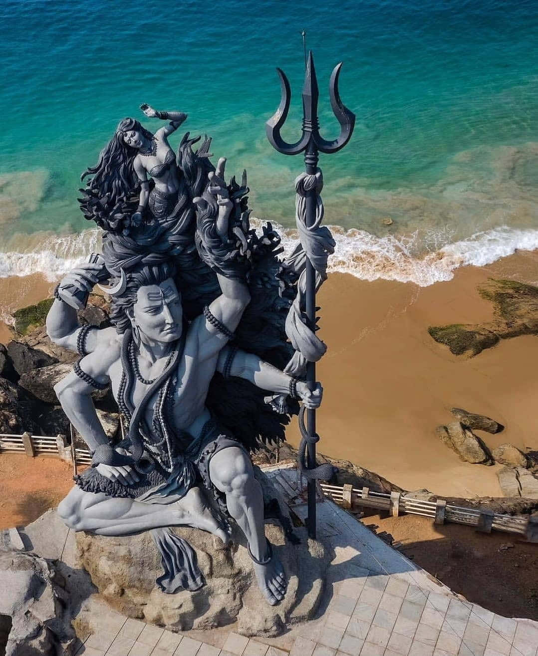 Kerala 桌布 壁紙 — 一座巨大的灰色石像濕婆，頭頂上有帕爾瓦蒂，手持三叉戟，矗立在岩石海邊懸崖上，俯瞰著碧綠色的海浪。