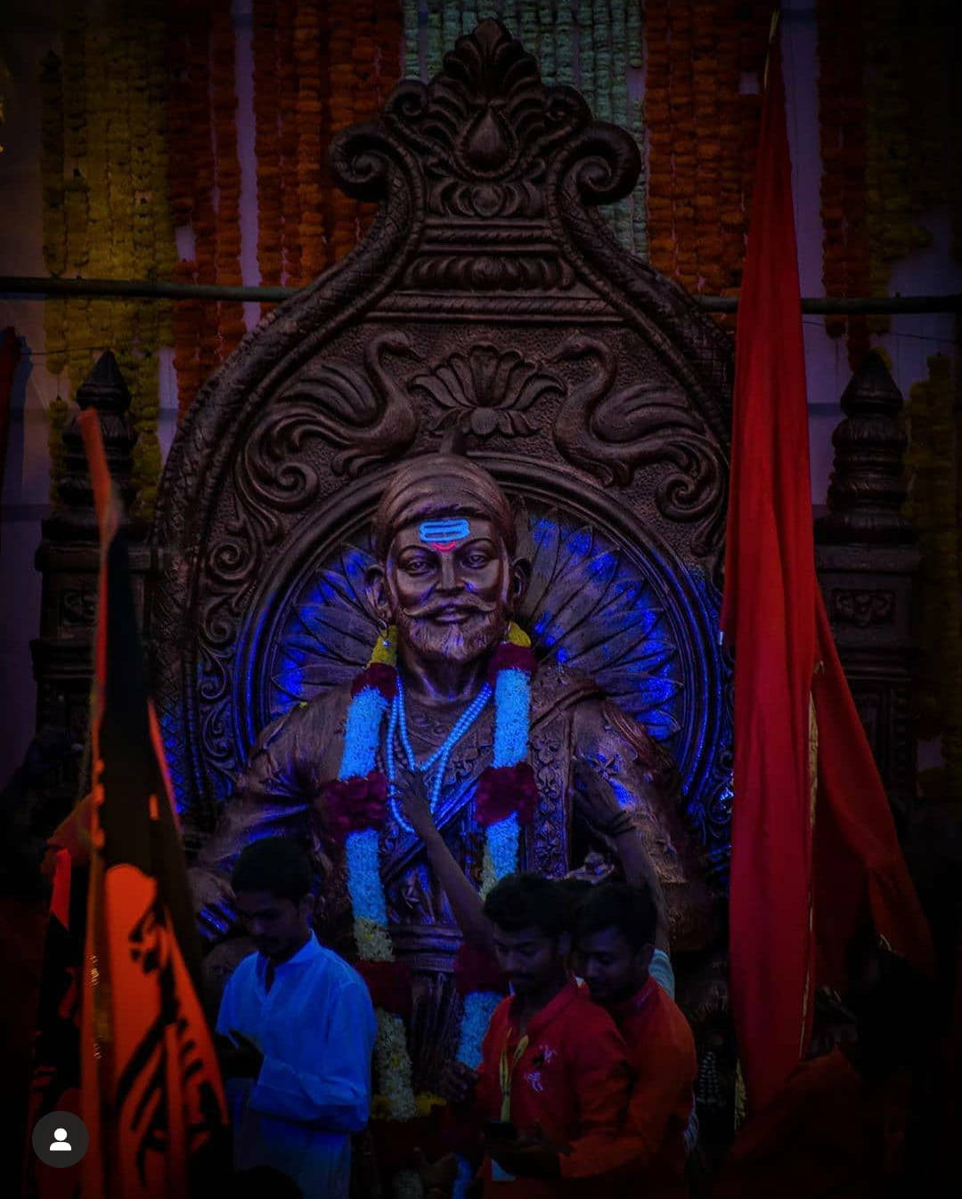  壁紙 — 一座巨大的照明神像，鬍鬚神祇裝飾著花環和紅旗，在寺廟環境中被信徒圍繞。