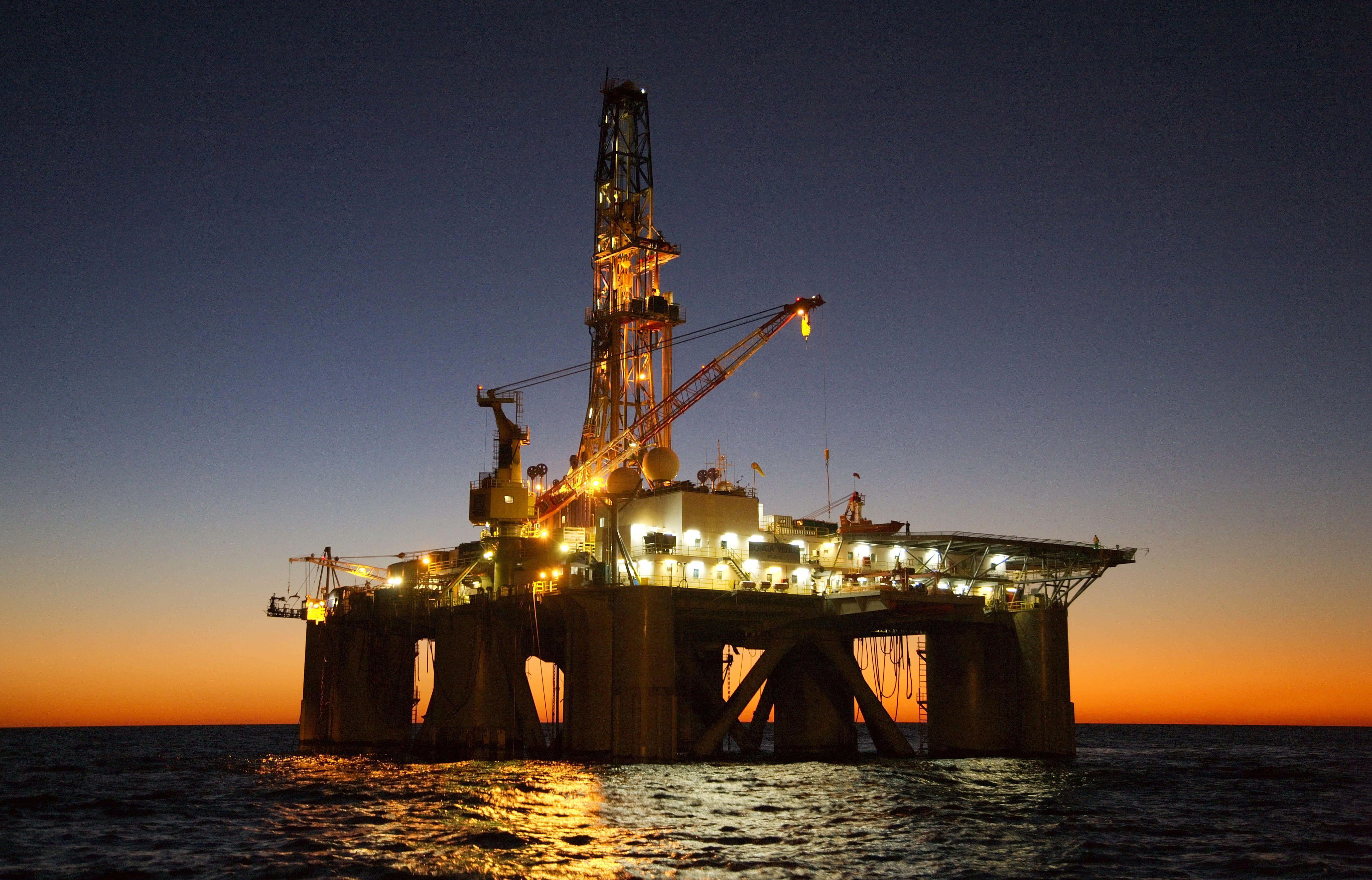Oil Rig 桌布 壁紙 — 一座巨大的照明離岸石油鑽井平台矗立在暮色天空中，橙色和藍色的漸變映照在平靜的海面上。
