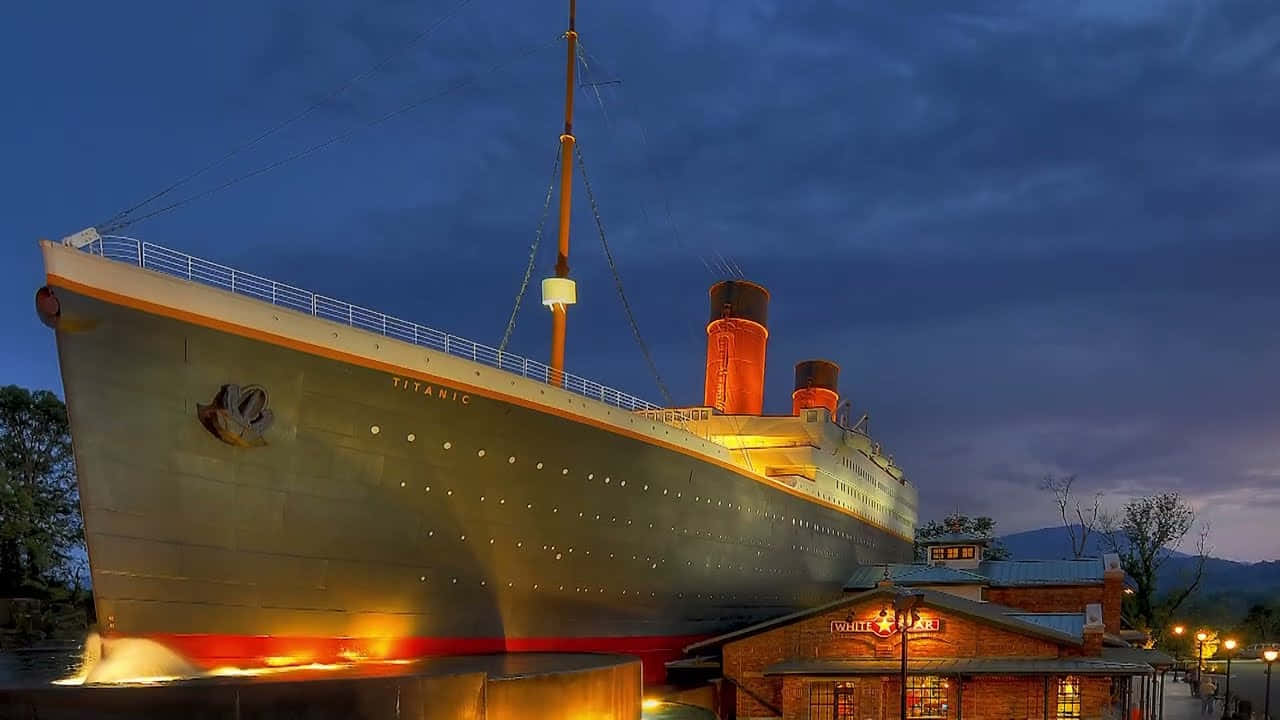 Titanic 桌布 壁紙 — 一座巨大的發光鐵達尼號複製品在黃昏時分雄偉地矗立，溫暖的燈光在深藍色的暮色天空中閃耀。