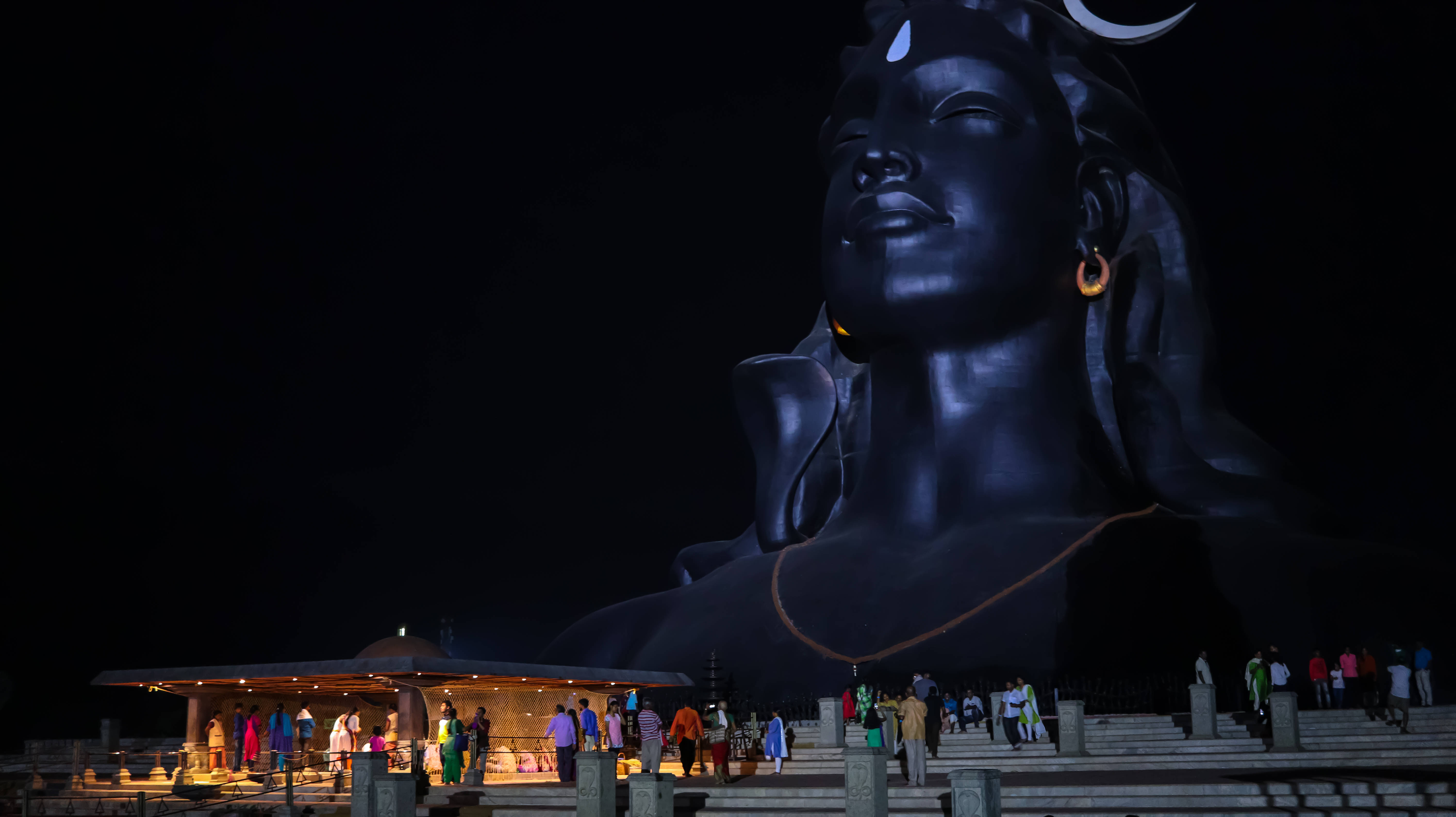 adiyogi shiva 桌布 壁紙 — 一座巨大的發光黑色濕婆神像，帶有新月和蛇，夜晚聳立在遊客上方。
