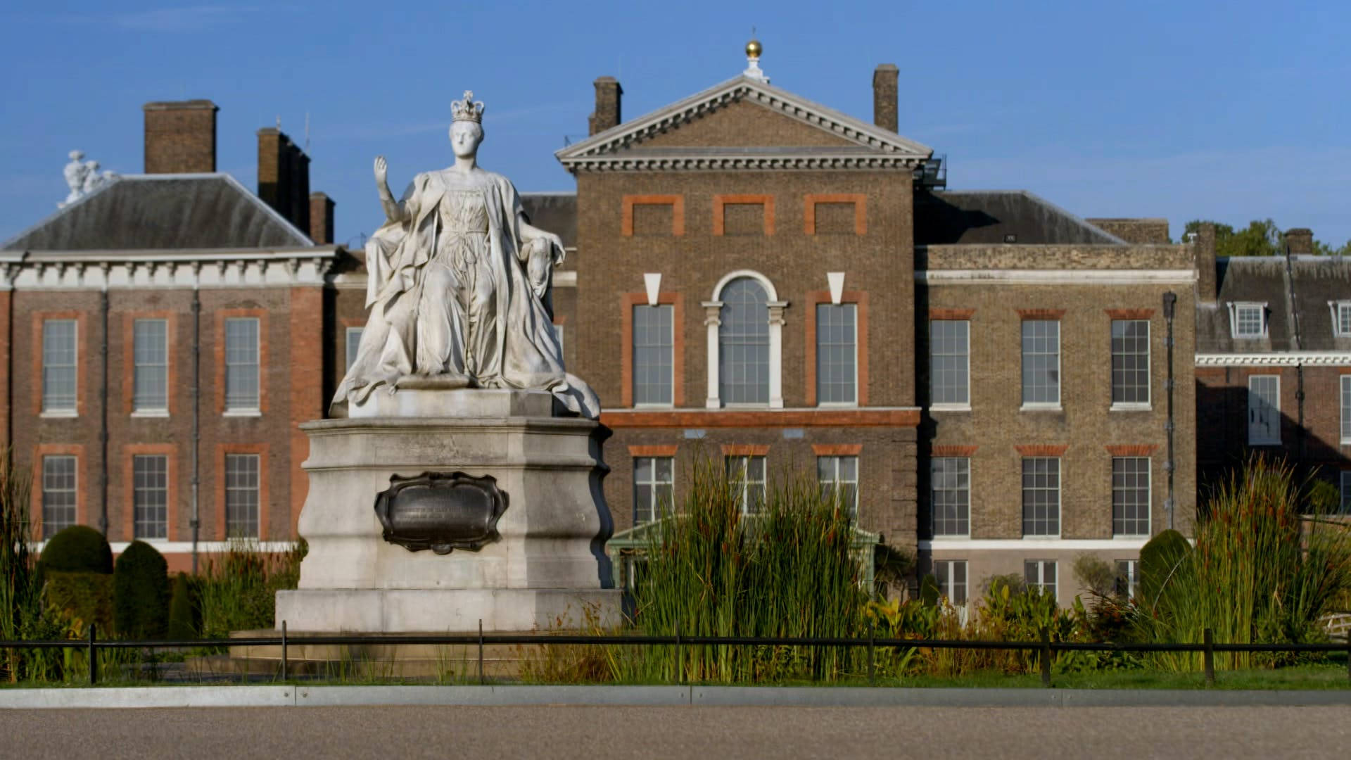 England 桌布 壁紙 — 一座巨大的白色大理石雕像，一位加冕的女王，矗立在歷史悠久的磚造宮殿前，背景是清澈的藍天。