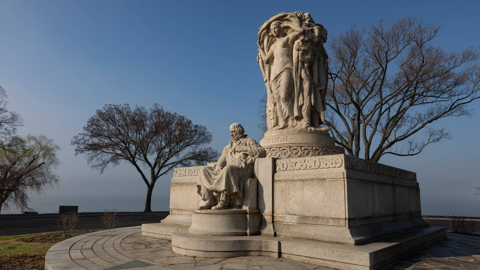 Statue Aesthetic 桌布 壁紙 — 一座巨大的石頭紀念碑，上面有坐著和站著的人物，矗立在晴朗的藍天下，周圍是光禿禿的樹木。