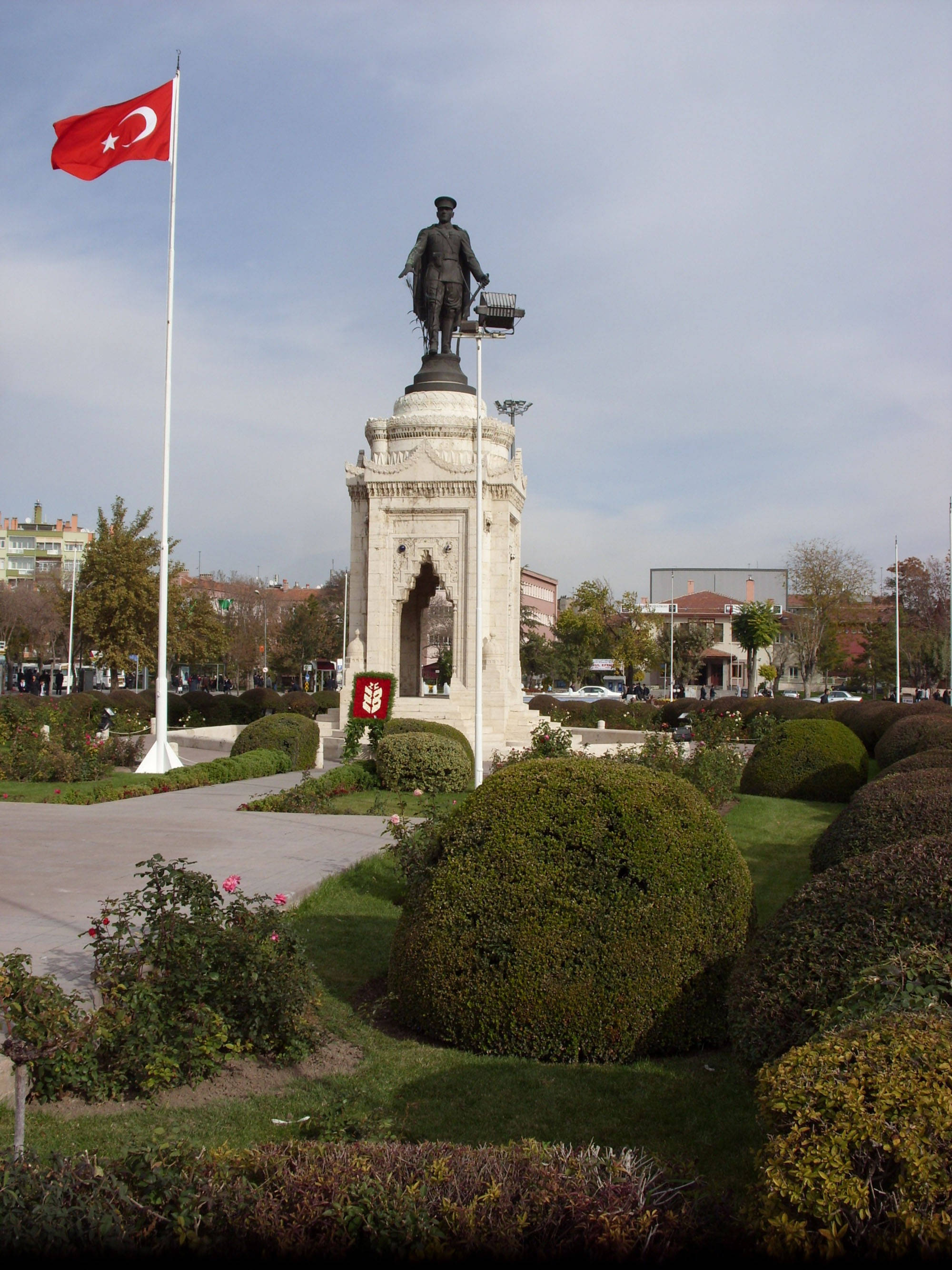 Konya 桌布 壁紙 — 一座巨大的石頭紀念碑頂部有一座銅像，矗立在修剪整齊的公園中，土耳其國旗飄揚，秋樹環繞。
