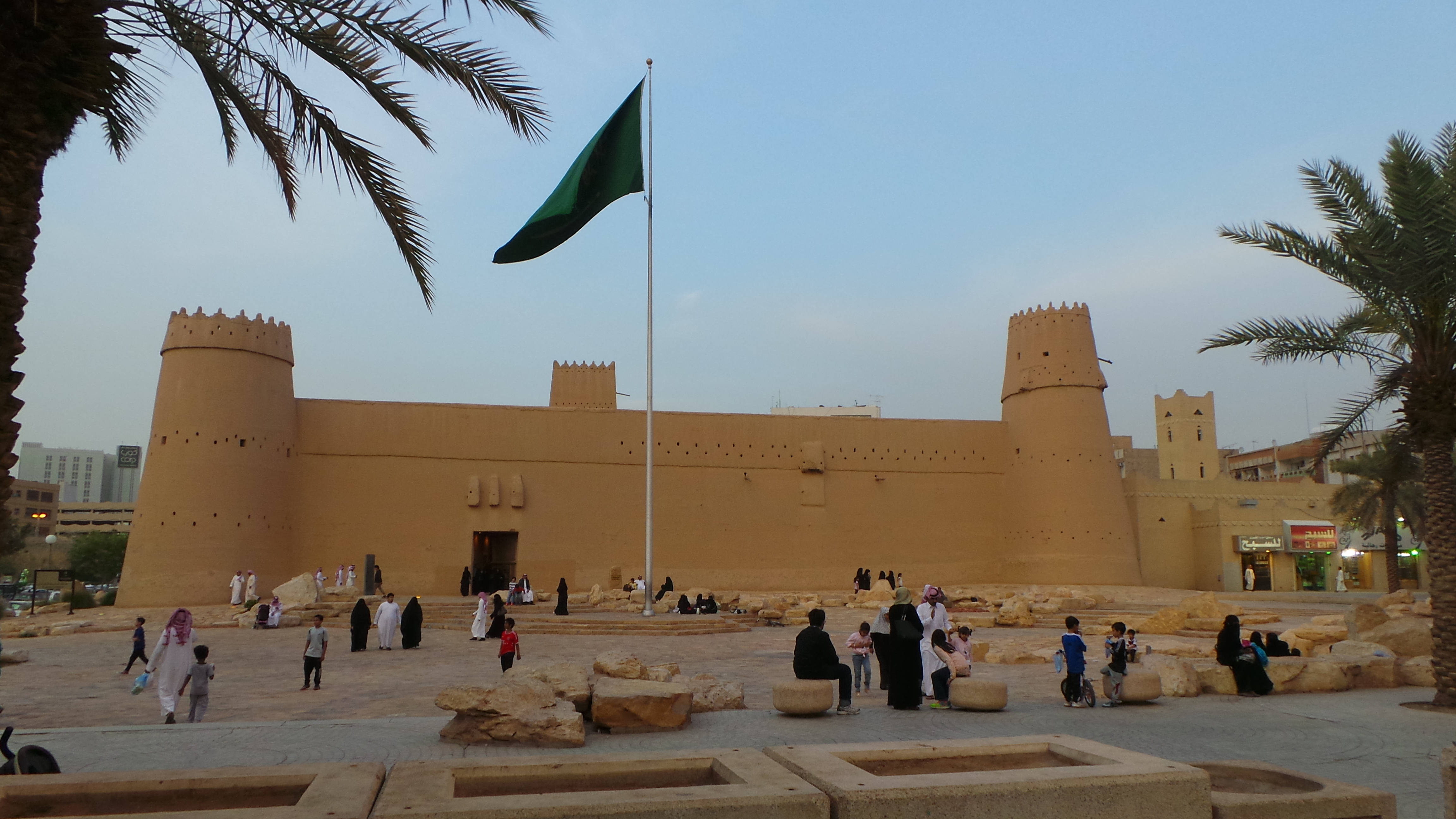 Saudi 桌布 壁紙 — 一座巨大的米色堡壘，帶有鋸齒狀塔樓，矗立在晴朗的天空下，人們在棕櫚樹和石凳間漫步或坐著。