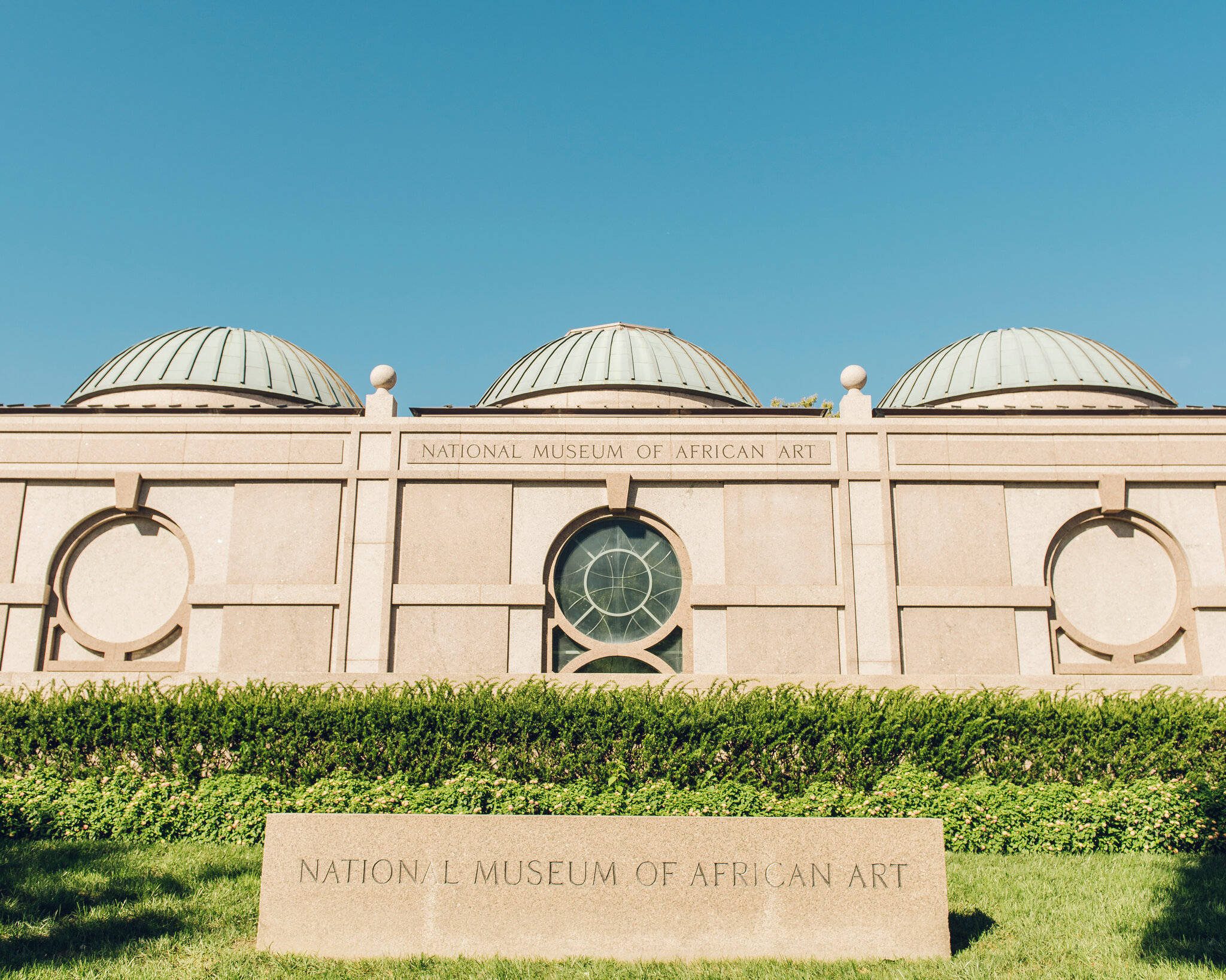 Smithsonian 桌布 壁紙 — 一座巨大的米色建築，有三個綠色圓頂和圓形窗戶，矗立在晴朗的藍天下，前面有修剪整齊的樹籬和石頭標誌。
