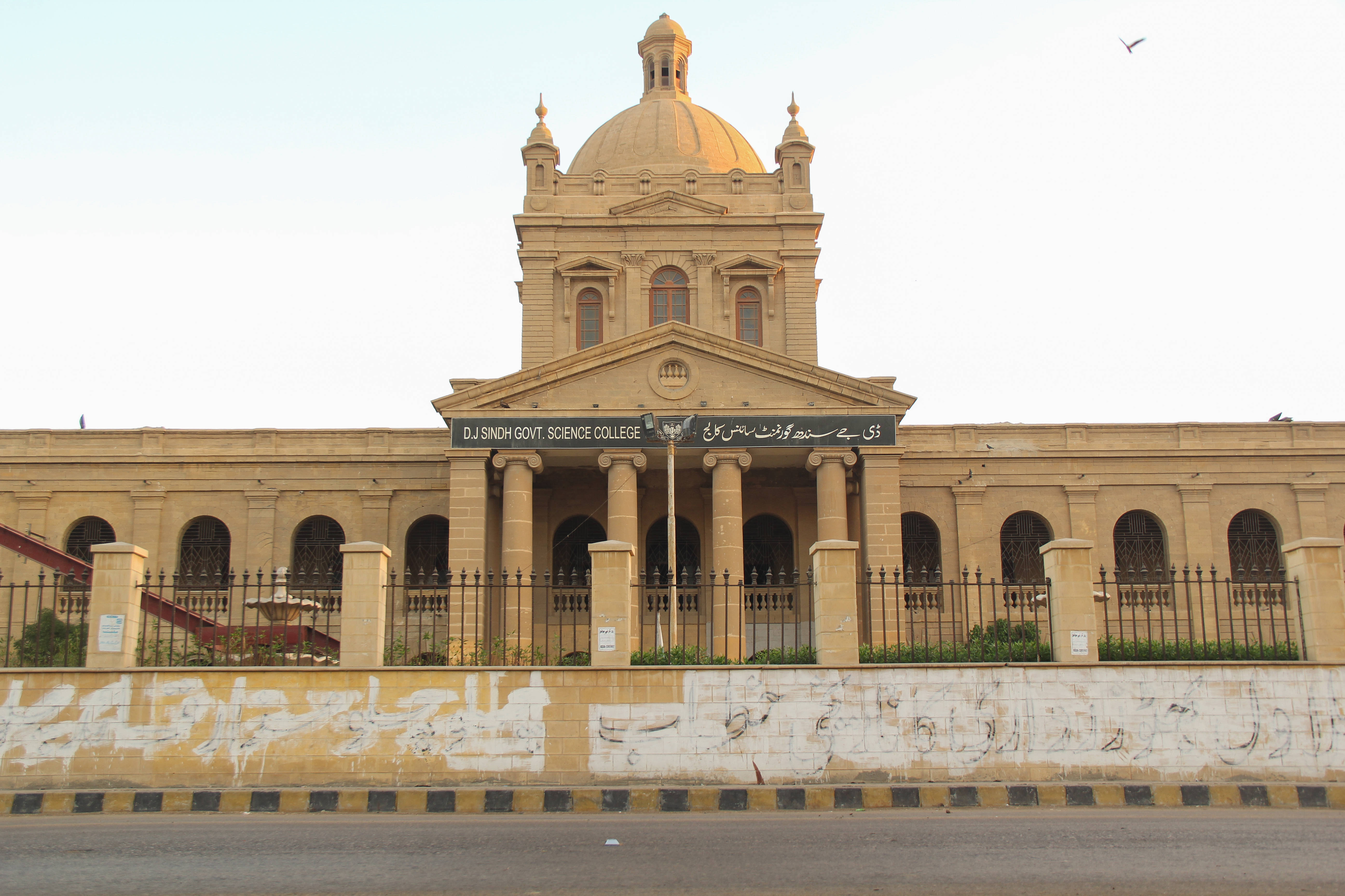 Karachi 桌布 壁紙 — 一座巨大的米色政府科學學院建築，中央有圓頂和柱廊，矗立在蒼白的天空下，前景牆上有塗鴉。