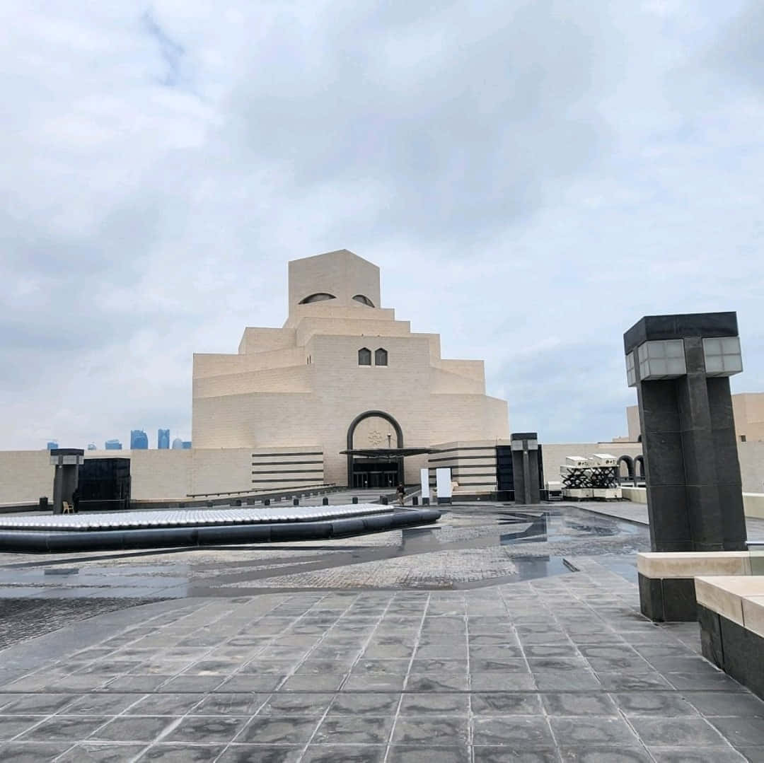 建築 桌布 壁紙 — 一座巨大的米色階梯式博物館建築，帶有拱形入口，矗立在陰天之下，潮濕的路面反射出其現代建築風格。