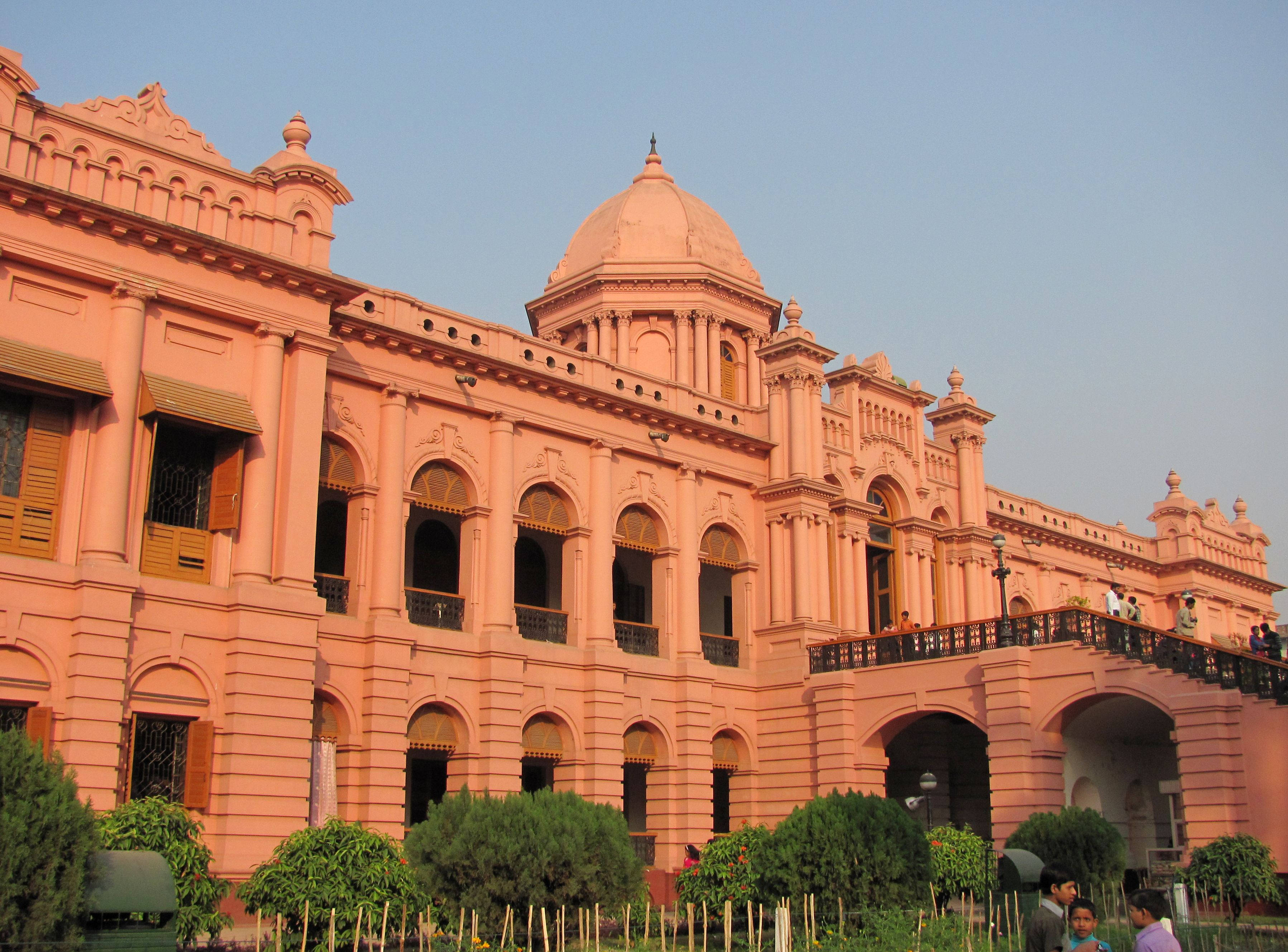 Dhaka 桌布 壁紙 — 一座巨大的粉紅色歷史建築，帶有圓頂和拱形窗戶，矗立在晴朗的藍天下，前景有綠色灌木。