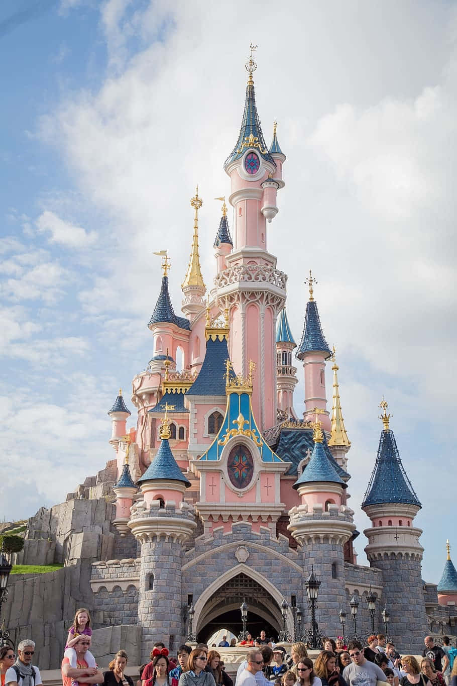 Disneyland Castle 桌布 壁紙 — 一座巨大的粉藍色城堡，金色尖塔聳立在部分多雲的天空下的人群之上。