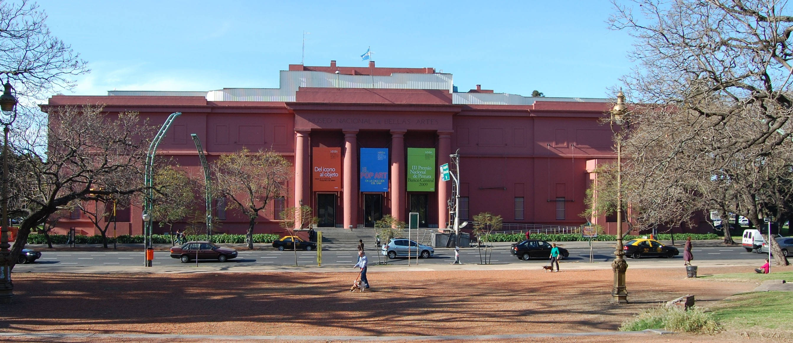 Aires 桌布 壁紙 — 一座巨大的紅磚博物館建築，帶有古典柱廊，矗立在清澈的藍天下，周圍是落葉樹木和鋪砌的廣場，廣場上有行人和汽車。