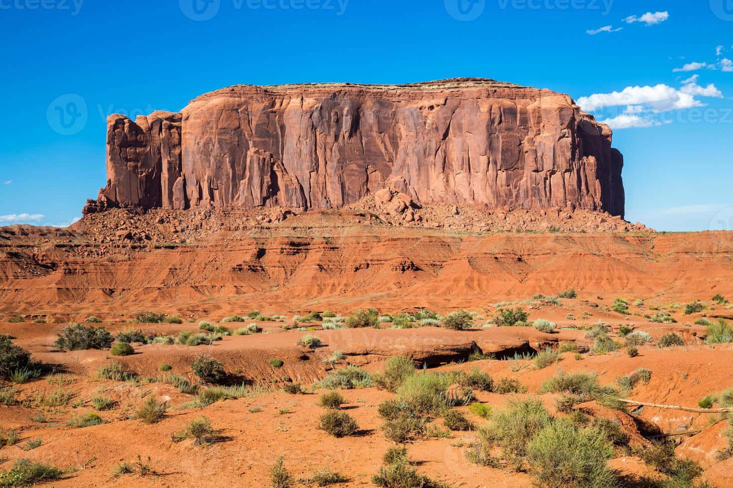 Arizona 桌布 壁紙 — 一座巨大的紅色砂岩孤峰在晴朗的藍天下戲劇性地聳立，俯瞰著崎嶇的沙漠景觀，點綴著稀疏的綠色灌木。