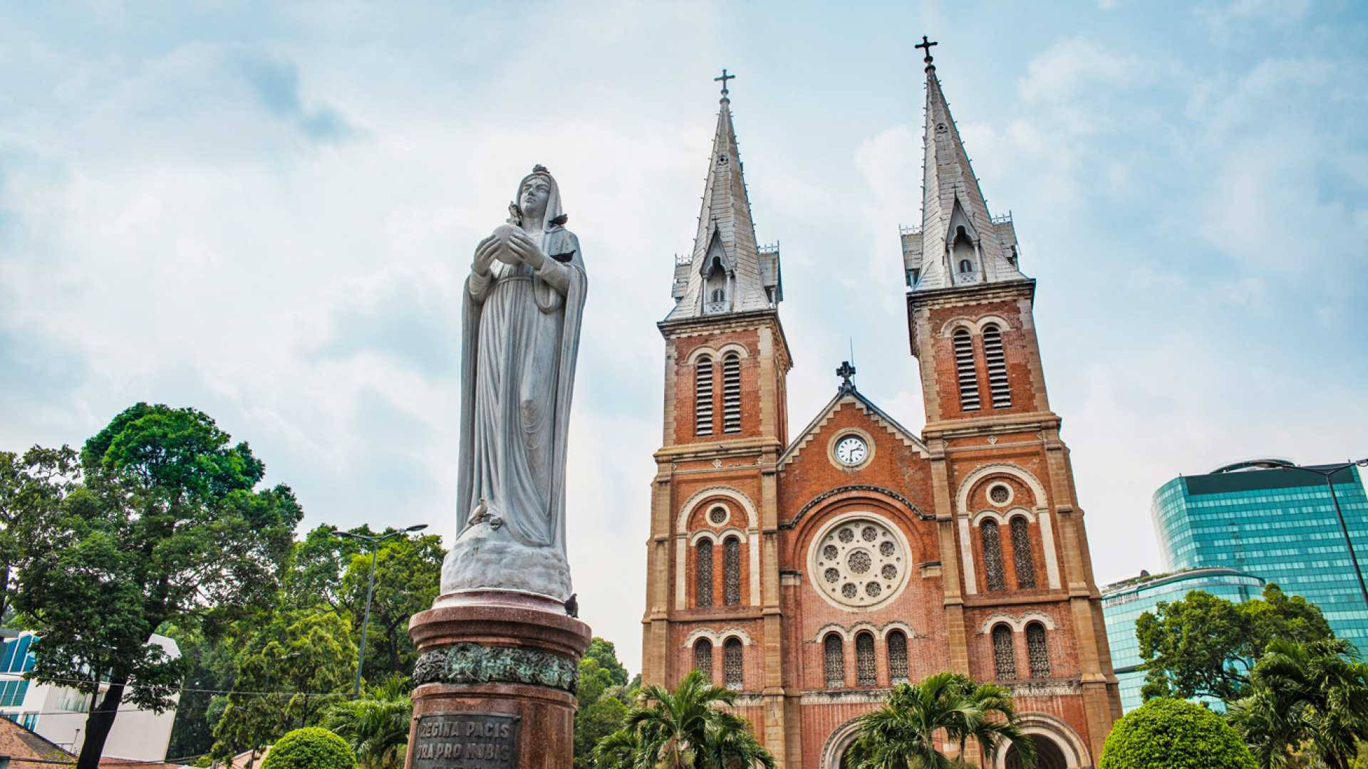 Ho Chi Minh City 桌布 壁紙 — 一座巨大的聖母瑪利亞石像矗立在紅磚哥德式大教堂前，雙塔尖頂在部分多雲的天空下。
