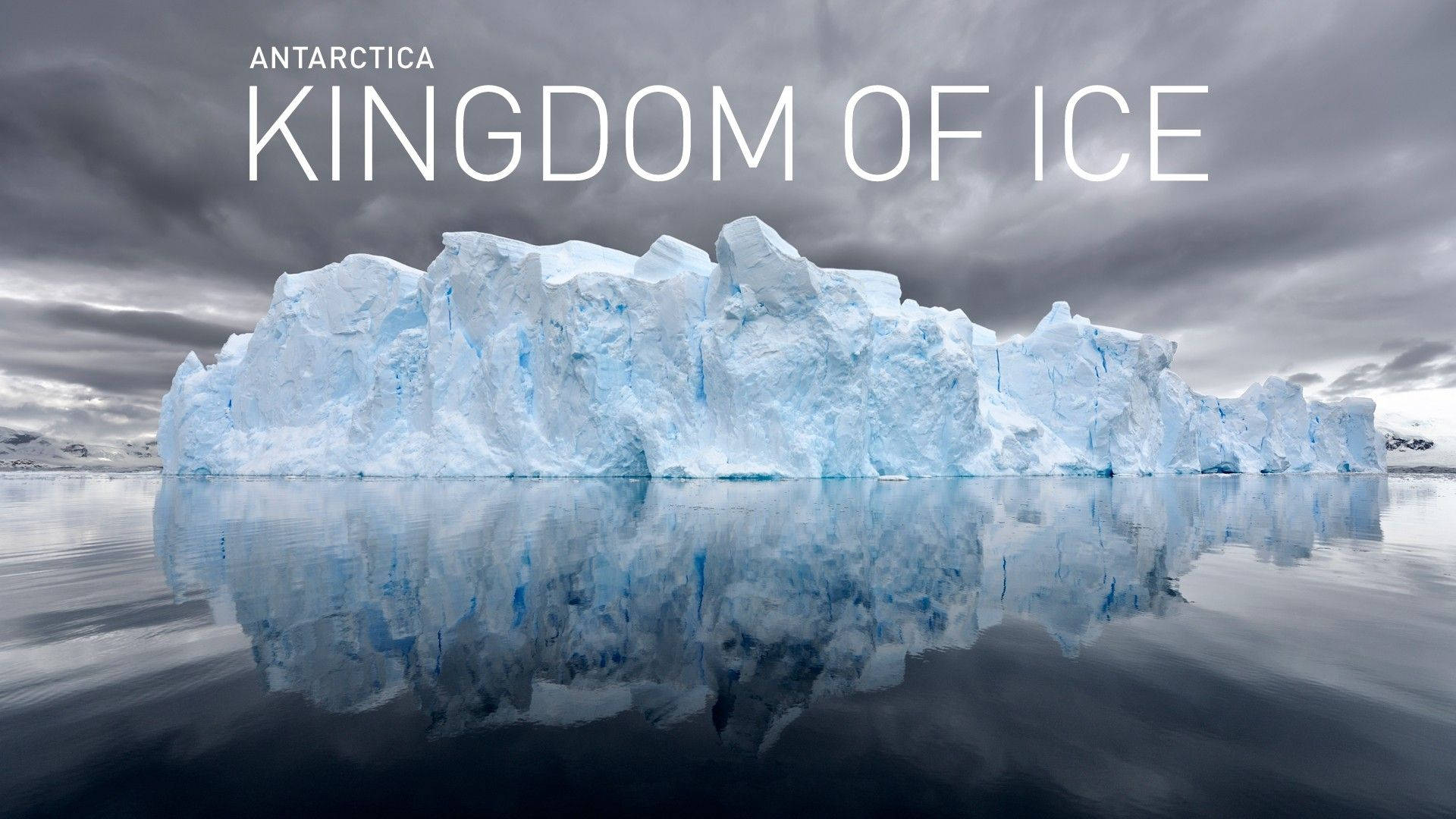 Antarctica 桌布 壁紙 — 一座巨大的藍色冰山在風暴天空下，於南極海域中平靜漂浮，並帶有完美倒影。
