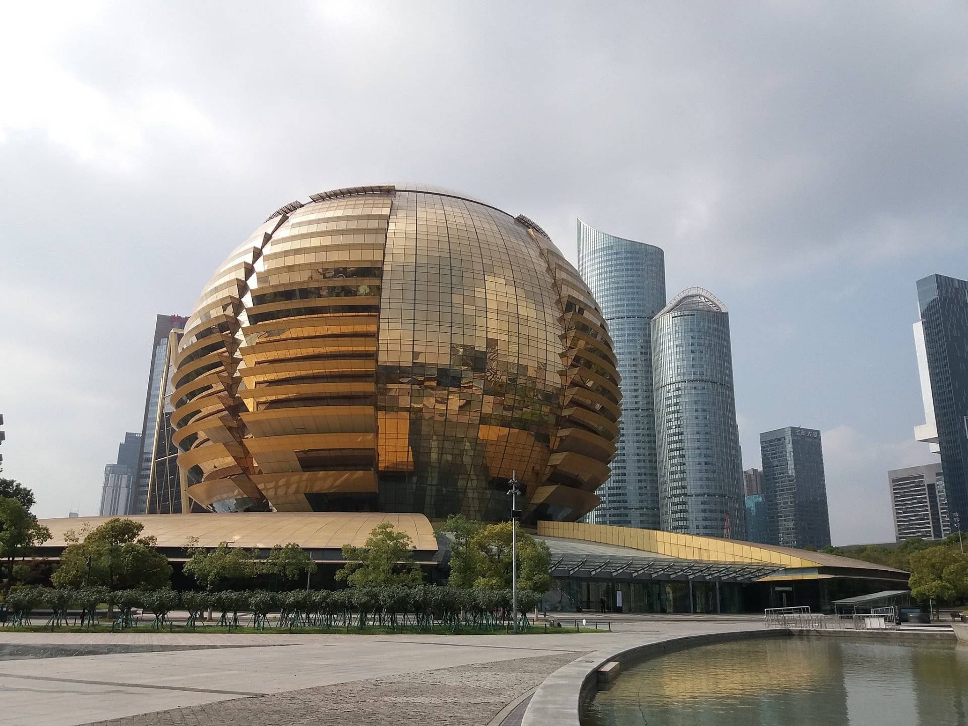 Hangzhou 桌布 壁紙 — 一座巨大的金色球形建築，帶有反光面板，在陰天天空下，矗立在現代城市景觀中，周圍環繞著高聳的玻璃摩天大樓。
