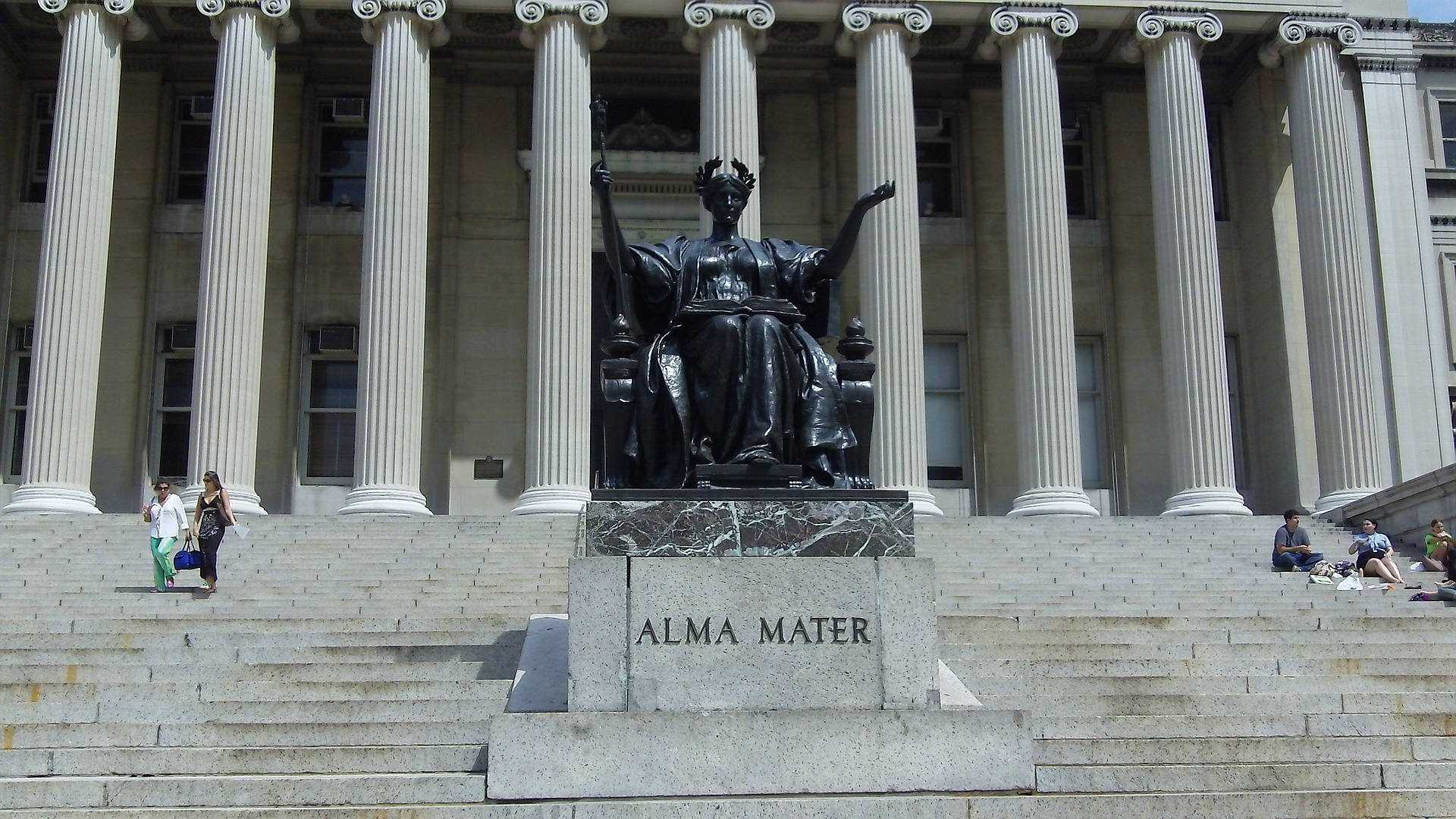 Library 桌布 壁紙 — 一座巨大的黑色Alma Mater雕像坐落在新古典主義建築前的大理石台階上，建築有高大的柱子，在明亮的日光下。