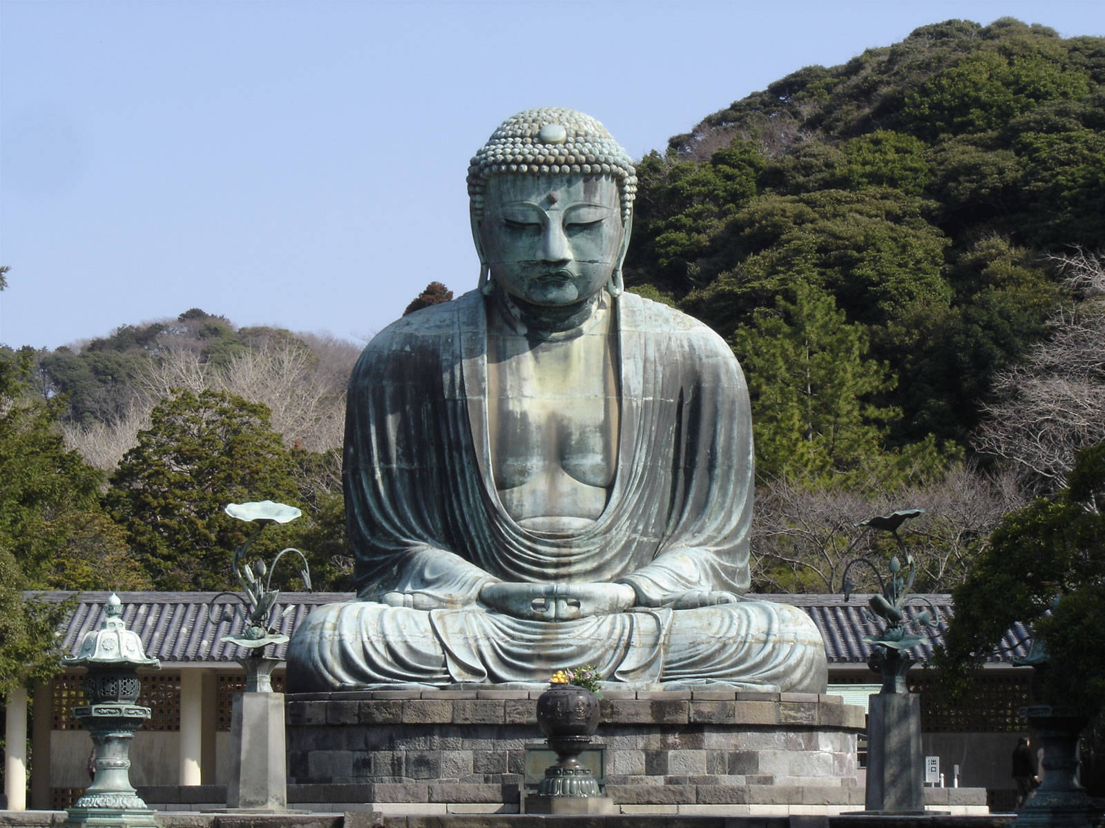 Statue Aesthetic 桌布 壁紙 — 一座巨大而寧靜的青銅佛像盤腿坐在石座上，周圍環繞著茂密的綠樹，頭頂是清澈的藍天。