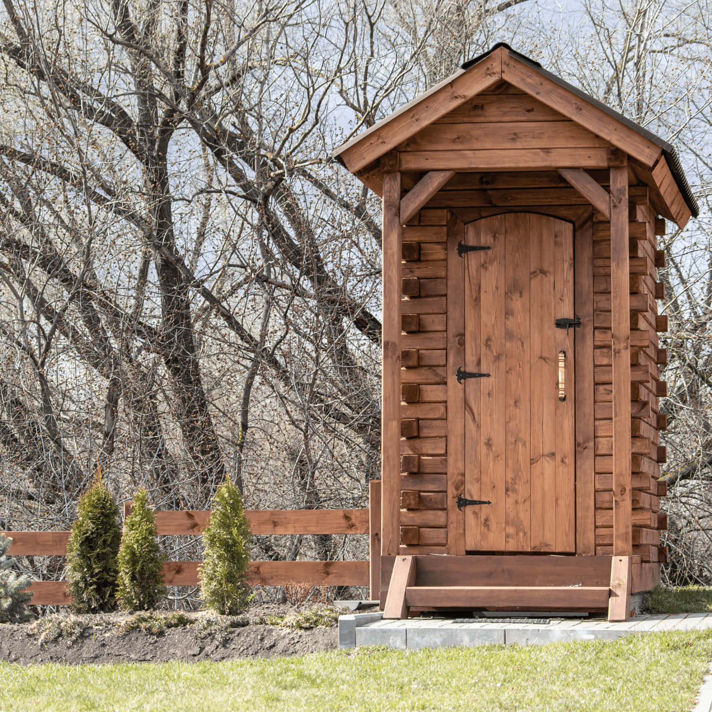 Outhouse 桌布 壁紙 — 一座帶有山牆屋頂的質樸木製戶外廁所，矗立在由光禿樹木和木柵欄圍繞的草地上。