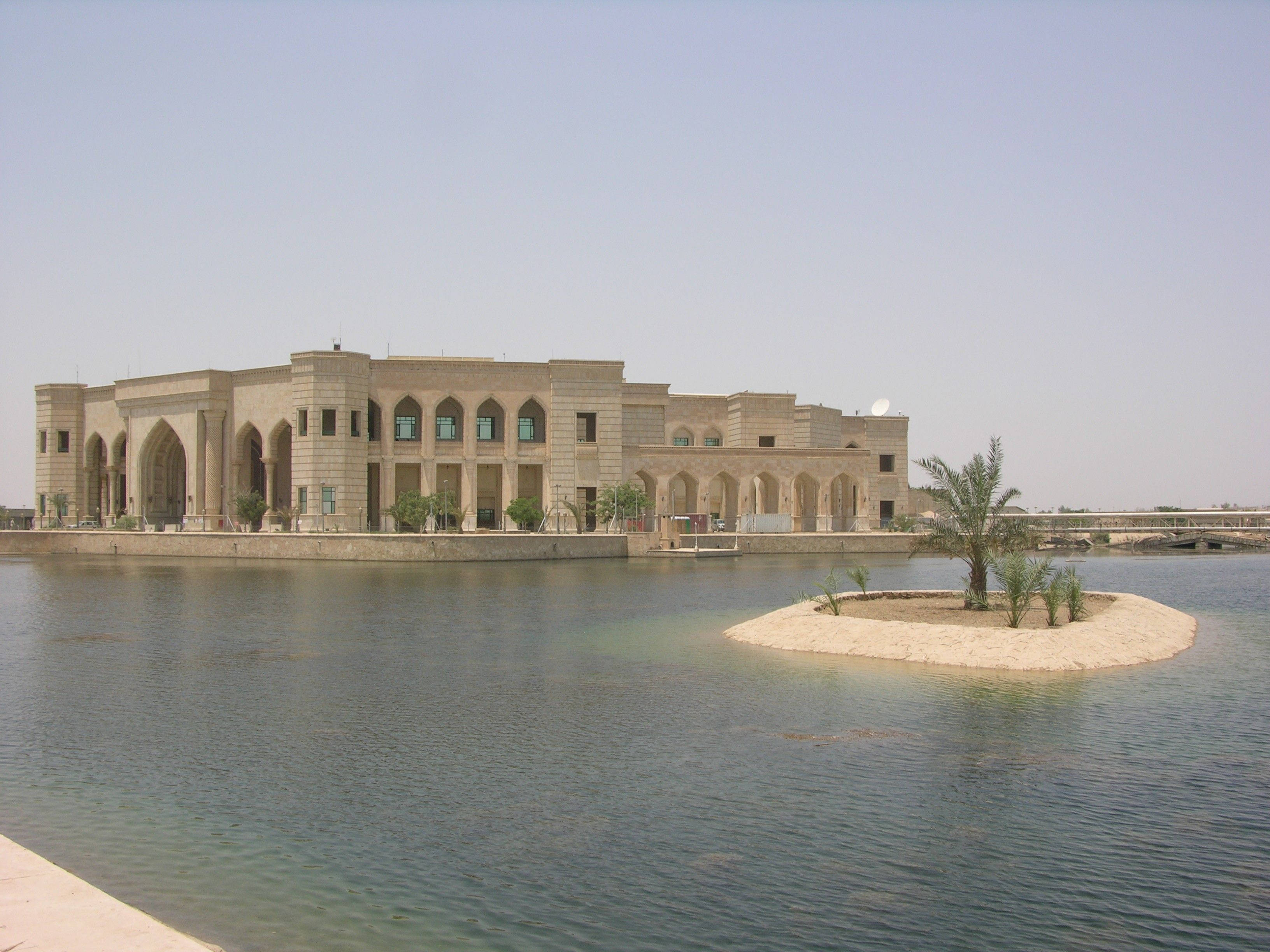 Iraq 桌布 壁紙 — 一座帶有拱形窗戶的大型米色石建築矗立在平靜的水邊，附近有一個小棕櫚樹島。