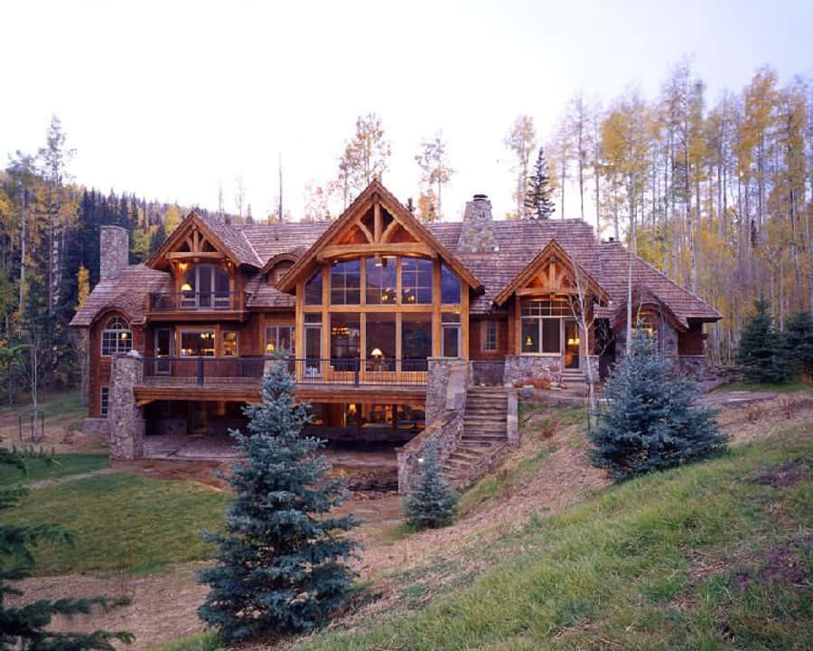 cabin 桌布 壁紙 — 一座帶有石頭裝飾和大窗戶的質樸木屋，坐落在長滿青草的山坡上，周圍環繞著秋葉樹木。