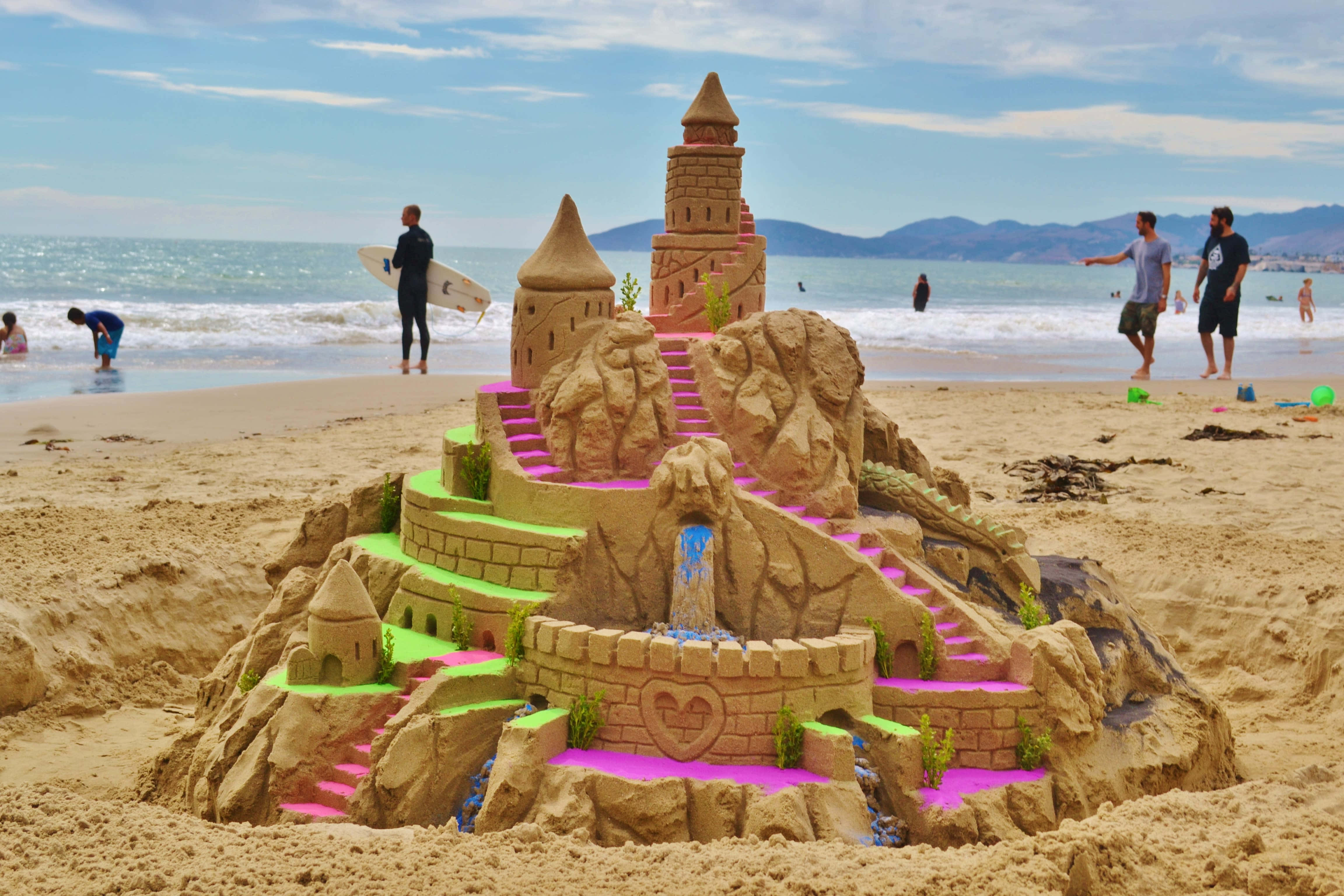 Sand Art 桌布 壁紙 — 一座帶有粉紅色和綠色點綴的鮮豔沙堡矗立在陽光明媚的海灘上，人們享受著海浪。