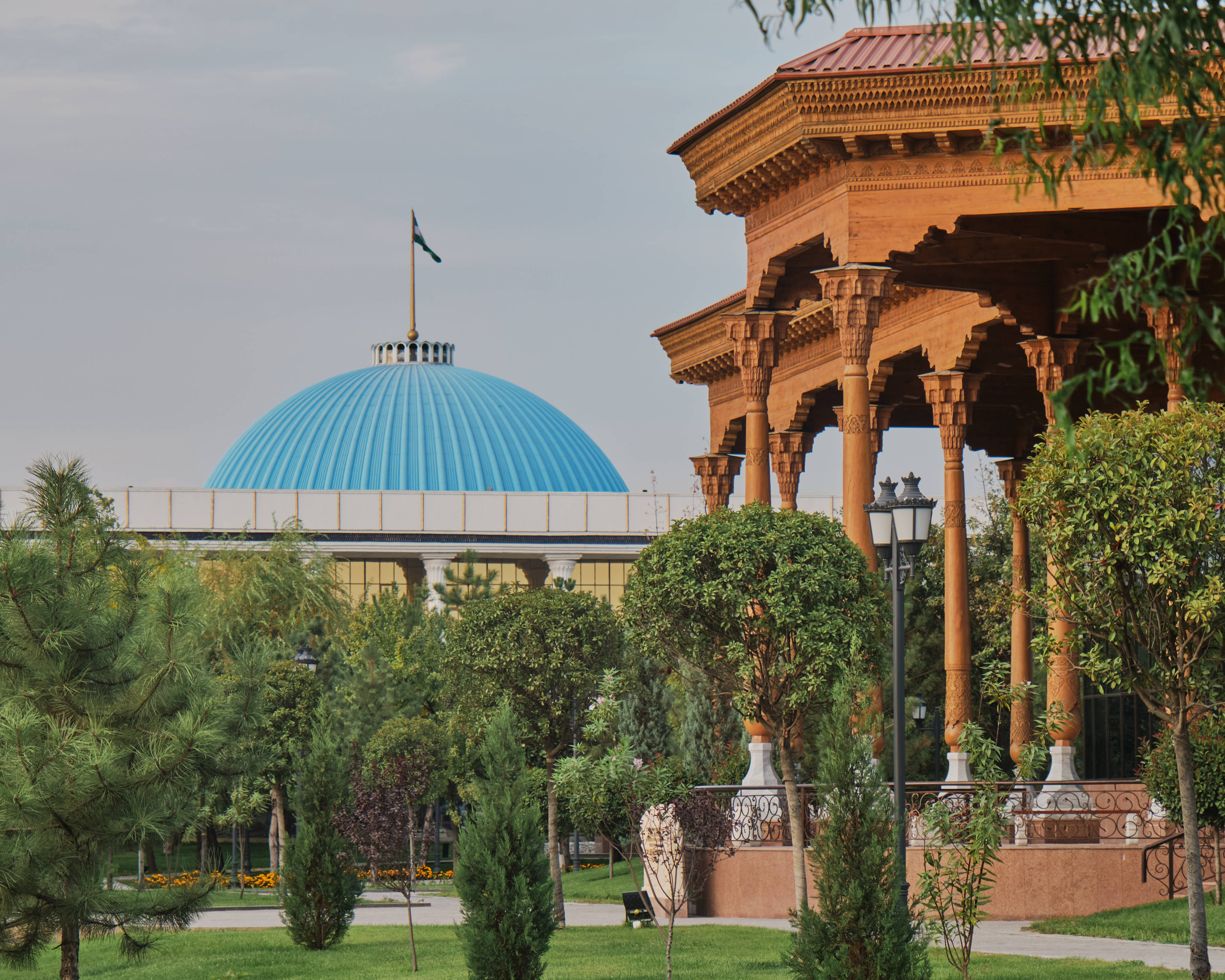 Uzbekistan 桌布 壁紙 — 一座帶有精緻雕刻的大型木製亭子矗立在鬱鬱蔥蔥的綠色公園中，樹木環繞，背景中有一座引人注目的藍色圓頂建築和旗幟，天空柔和。