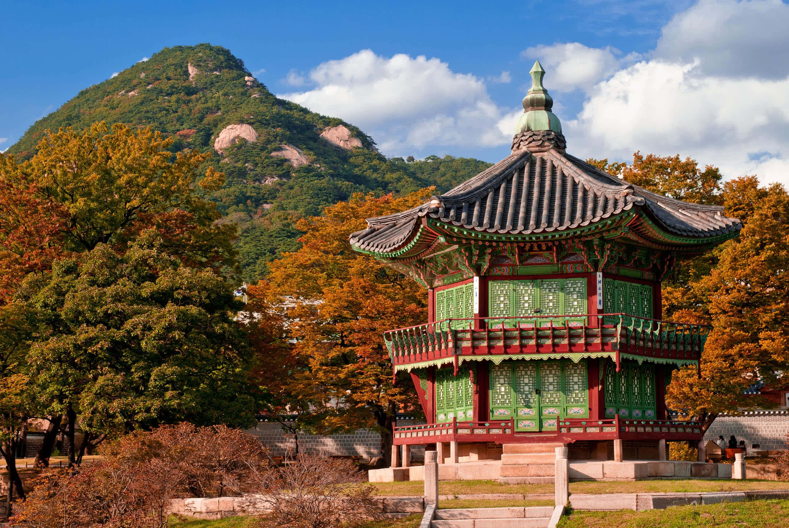 Architecture 桌布 壁紙 — 一座帶有綠色和紅色點綴的傳統韓國亭子矗立在秋樹之中，背景是森林覆蓋的山脈，藍天白雲。