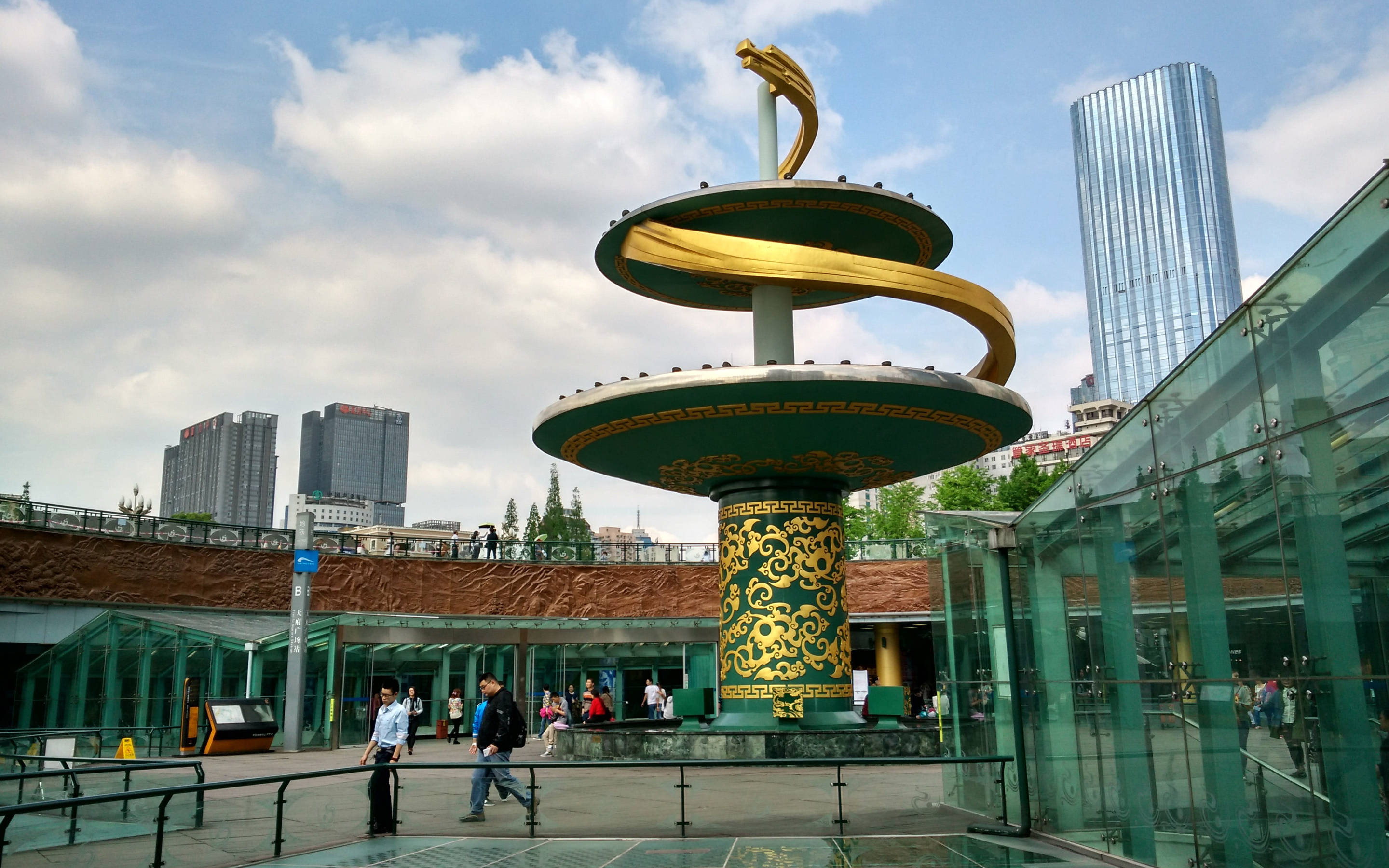 Chengdu 桌布 壁紙 — 一座帶有綠色點綴的金色螺旋雕塑矗立在現代城市廣場上，天空多雲，附近有玻璃建築和行人。