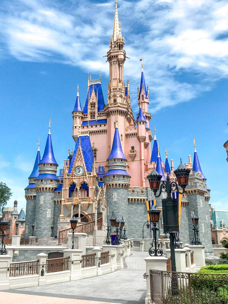 Disney Castle 桌布 壁紙 — 一座帶有藍色尖頂的宏偉粉紅色城堡，在晴朗藍天和白雲下高聳入雲。