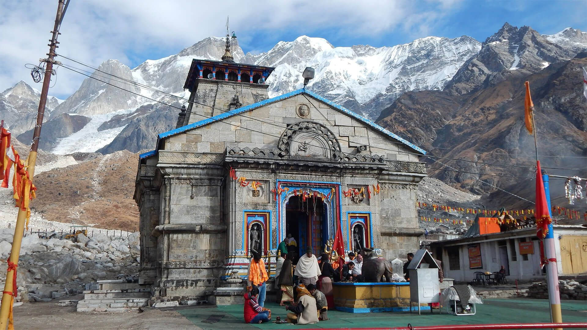 Kedarnath 桌布 壁紙 — 一座帶有藍色飾邊的石廟矗立在部分多雲的天空下，面對著白雪覆蓋的喜馬拉雅山峰。