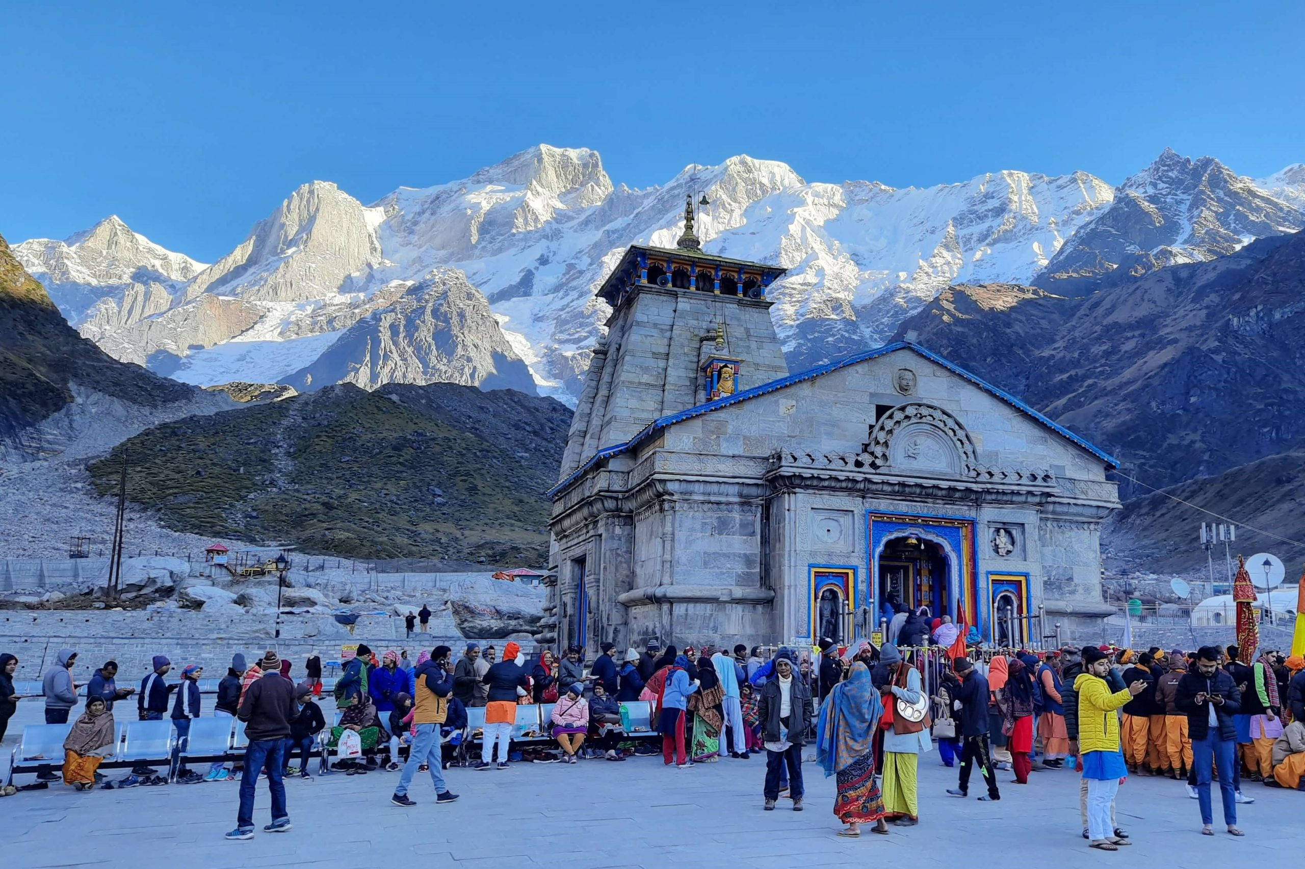Kedarnath 桌布 壁紙 — 一座帶有藍色點綴的鮮豔寺廟矗立在白雪覆蓋的喜馬拉雅山峰前，天空清澈湛藍，朝聖者身著色彩繽紛的服飾聚集於此。
