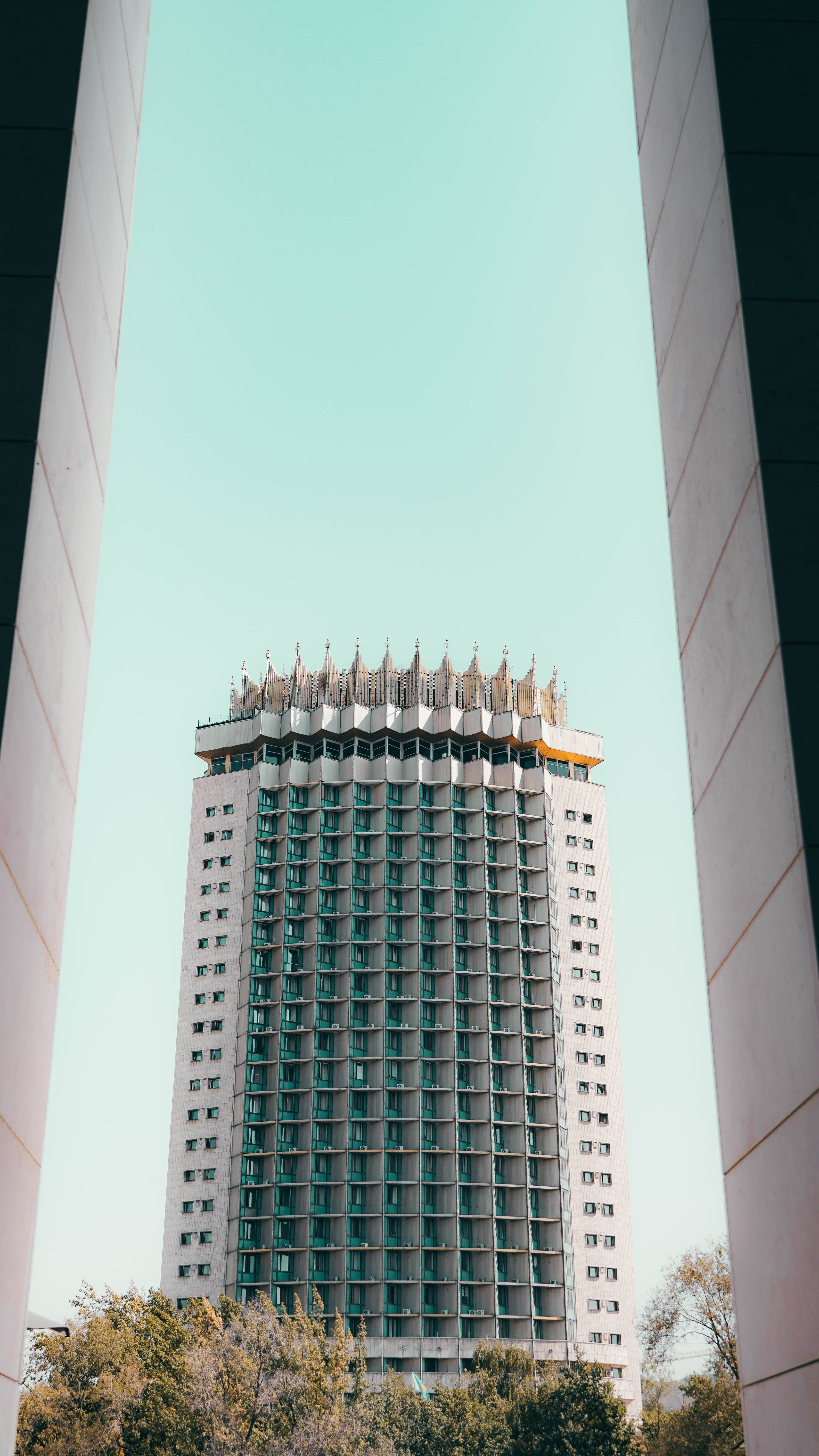 Kazakhstan 桌布 壁紙 — 一座帶有裝飾性冠頂的高層現代主義建築，在清澈淡藍的天空下，矗立於兩根混凝土柱之間。