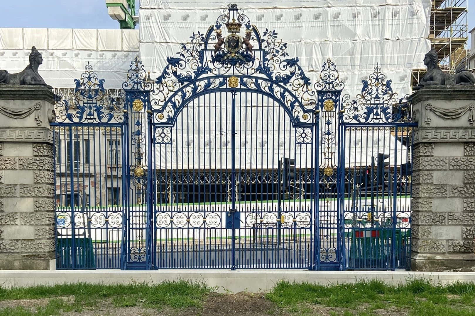 gate 桌布 壁紙 — 一座帶有金色點綴和石柱的華麗藍色鍛鐵大門，矗立在白色鷹架下的建築物前。