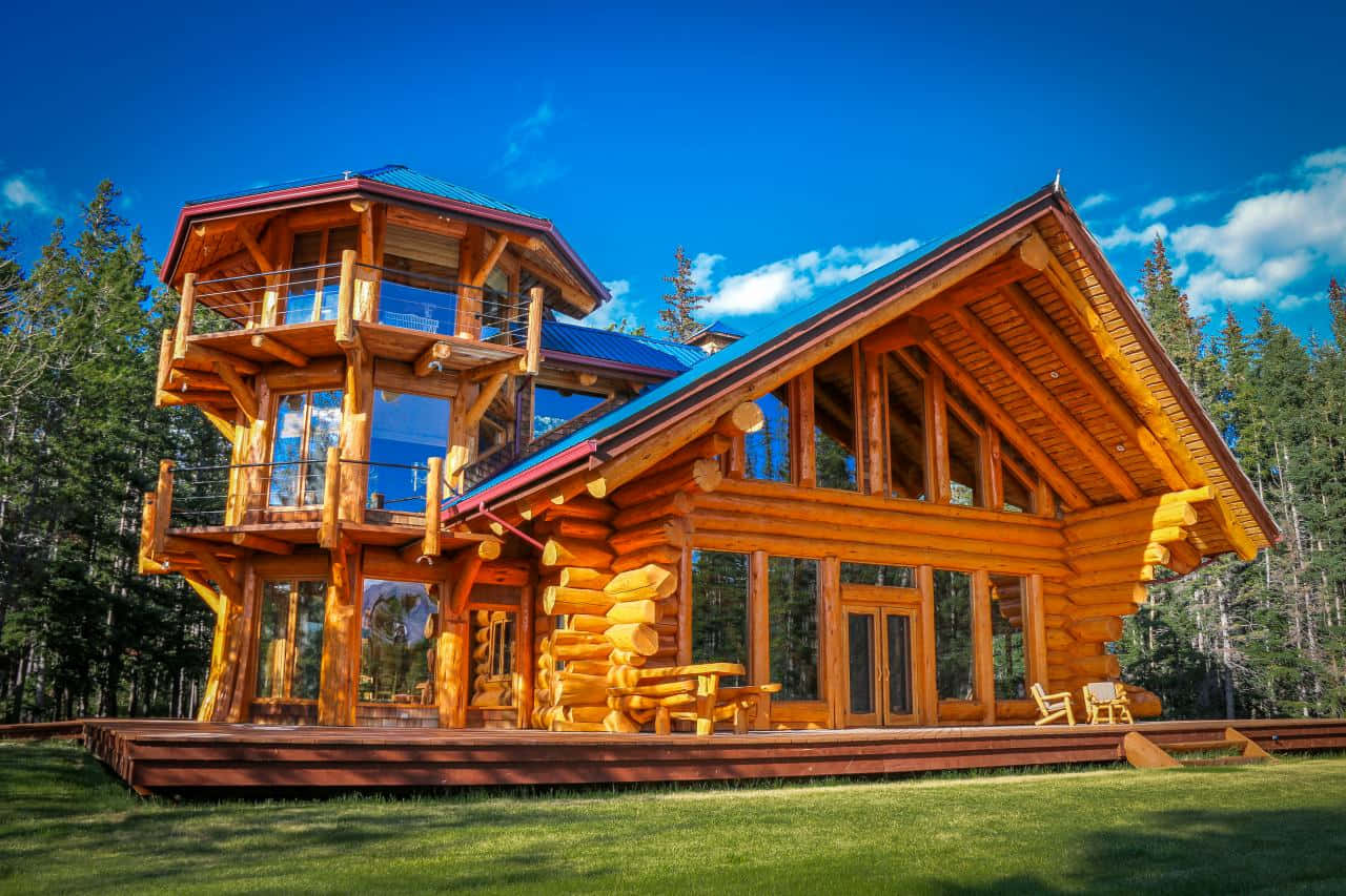 Log Cabin 桌布 壁紙 — 一座帶玻璃牆和陽台的大型木屋矗立在草地上，周圍是高聳的松樹，頭頂是明亮的藍天。