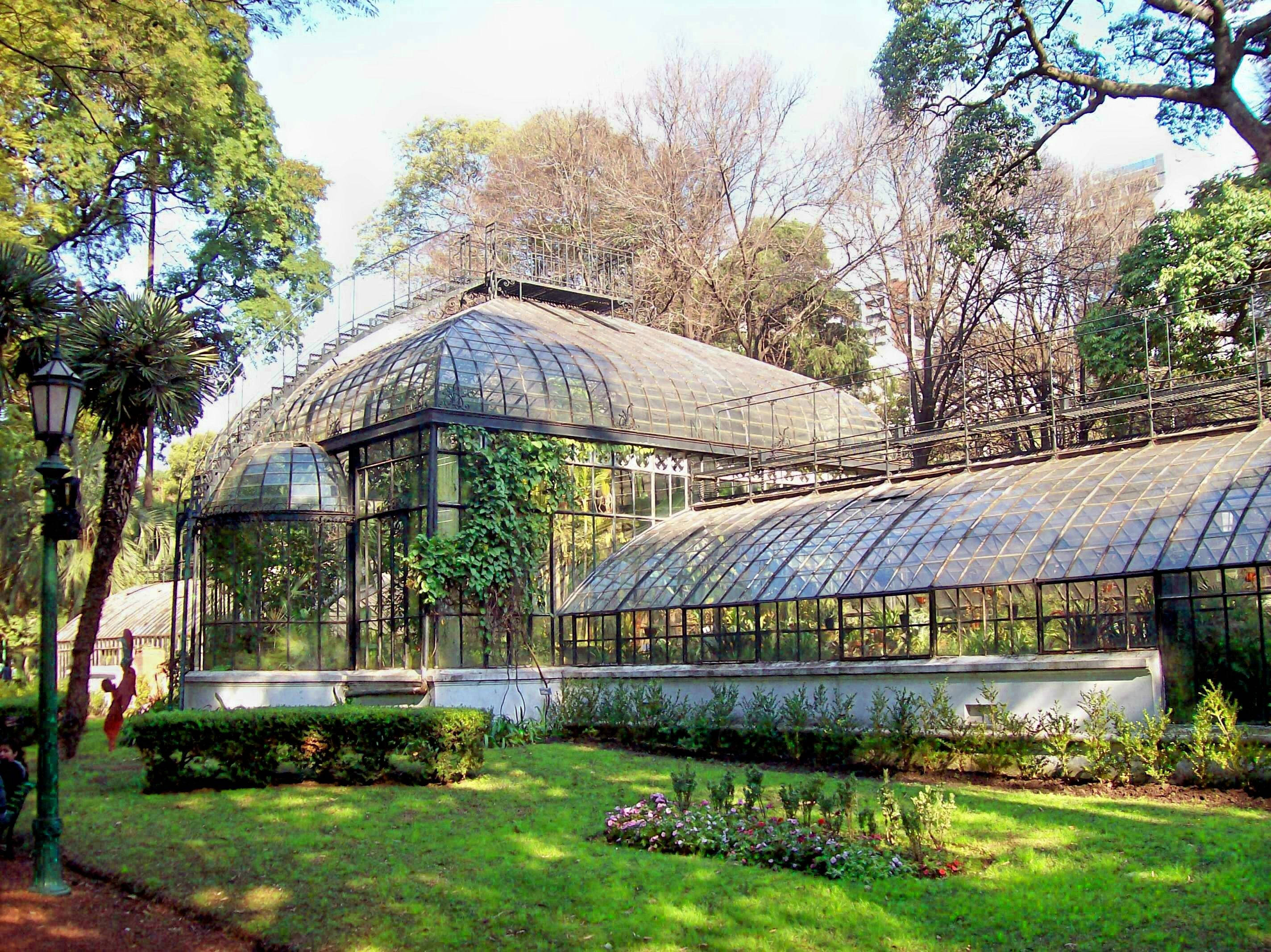 Aires 桌布 壁紙 — 一座帶鐵框的大型玻璃溫室矗立在陽光明媚的公園中，周圍環繞著茂密的綠植和盛開的花朵。
