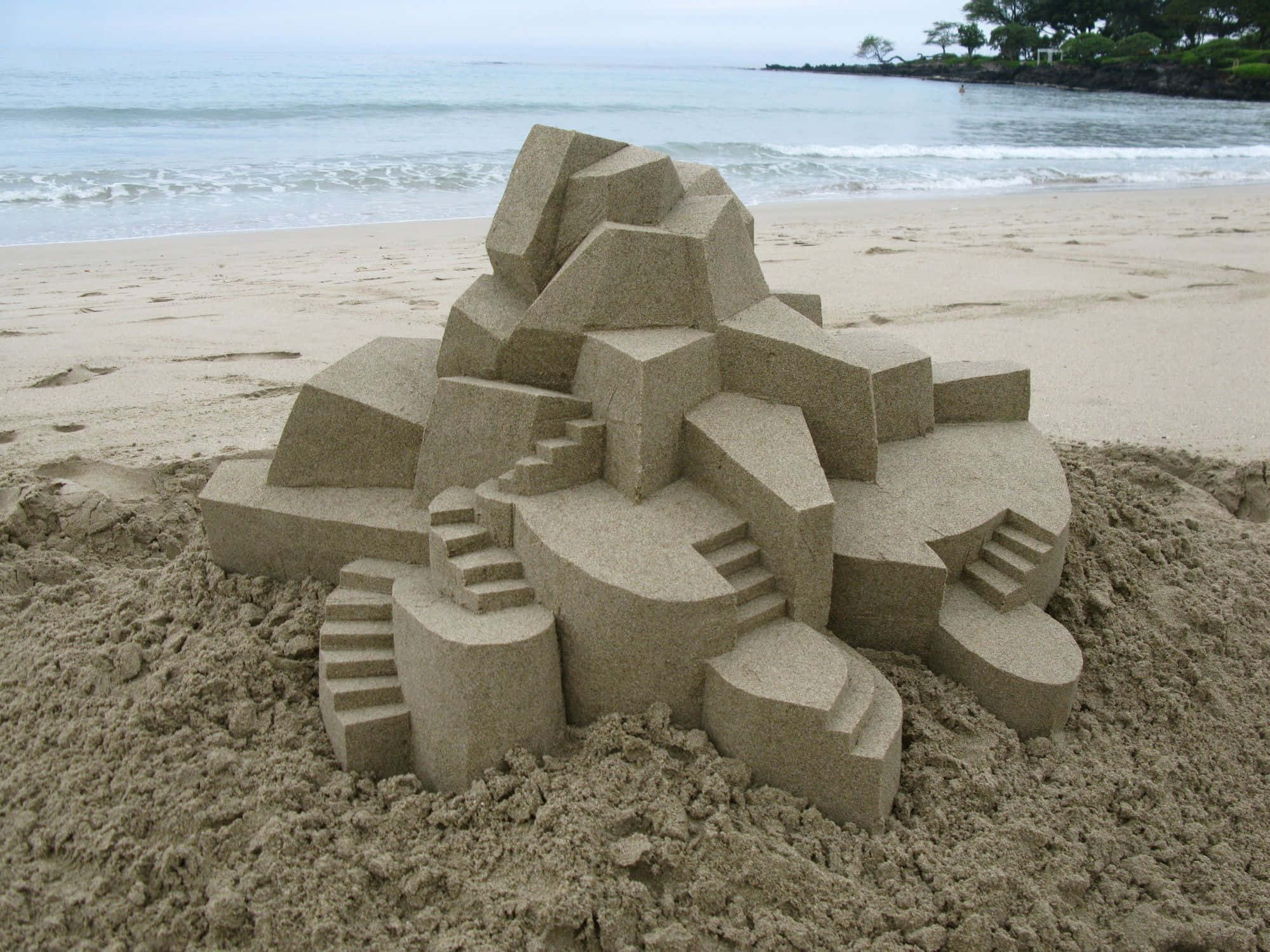 Sandcastles 桌布 壁紙 — 一座幾何沙堡，帶有棱角分明的塔樓和樓梯，矗立在沙灘上，背景有海浪和遠處的樹木，天空多雲。
