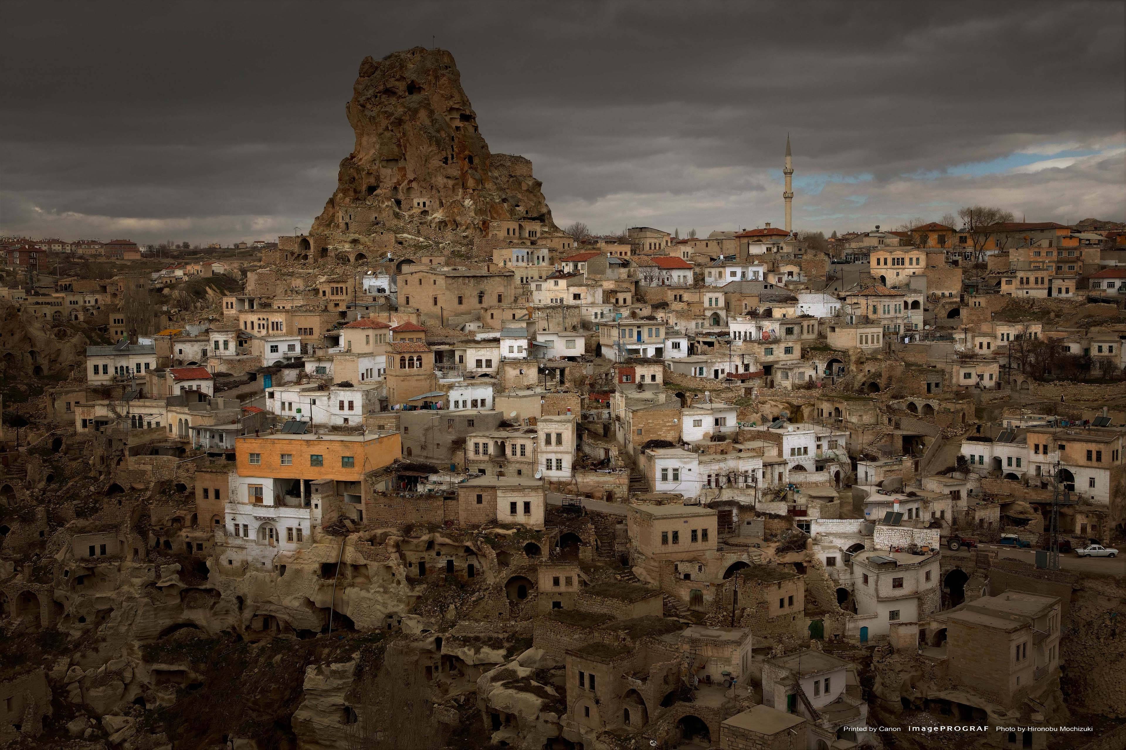 Cappadocia 桌布 壁紙 — 一座廣闊的古代洞穴城市依附在崎嶇的懸崖上，在沉重的暴風雨天空下，一座孤獨的宣禮塔刺穿雲層。