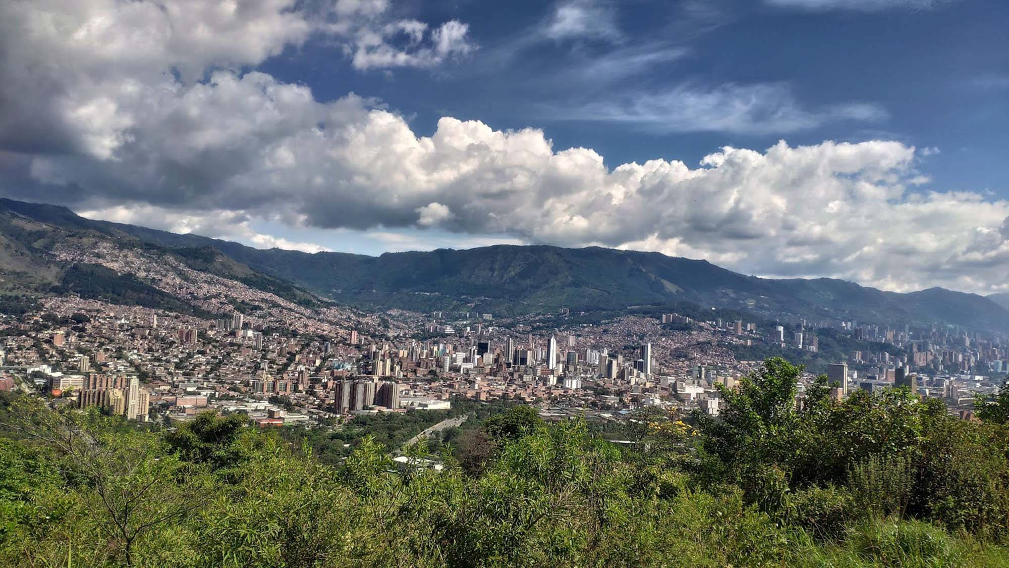 Medellin 桌布 壁紙 — 一座廣闊的城市依附在山坡上，天空戲劇性，充滿蓬鬆的白雲。