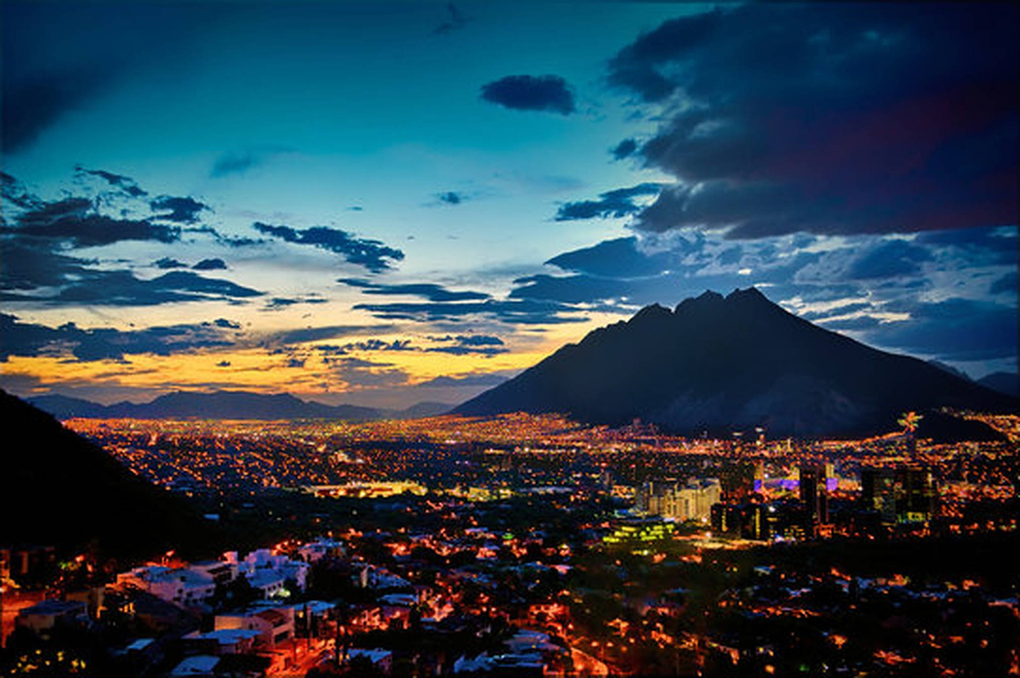 Monterrey 桌布 壁紙 — 一座廣闊的城市在戲劇性的暮色天空下亮起，遠處有黑暗的山脈輪廓。