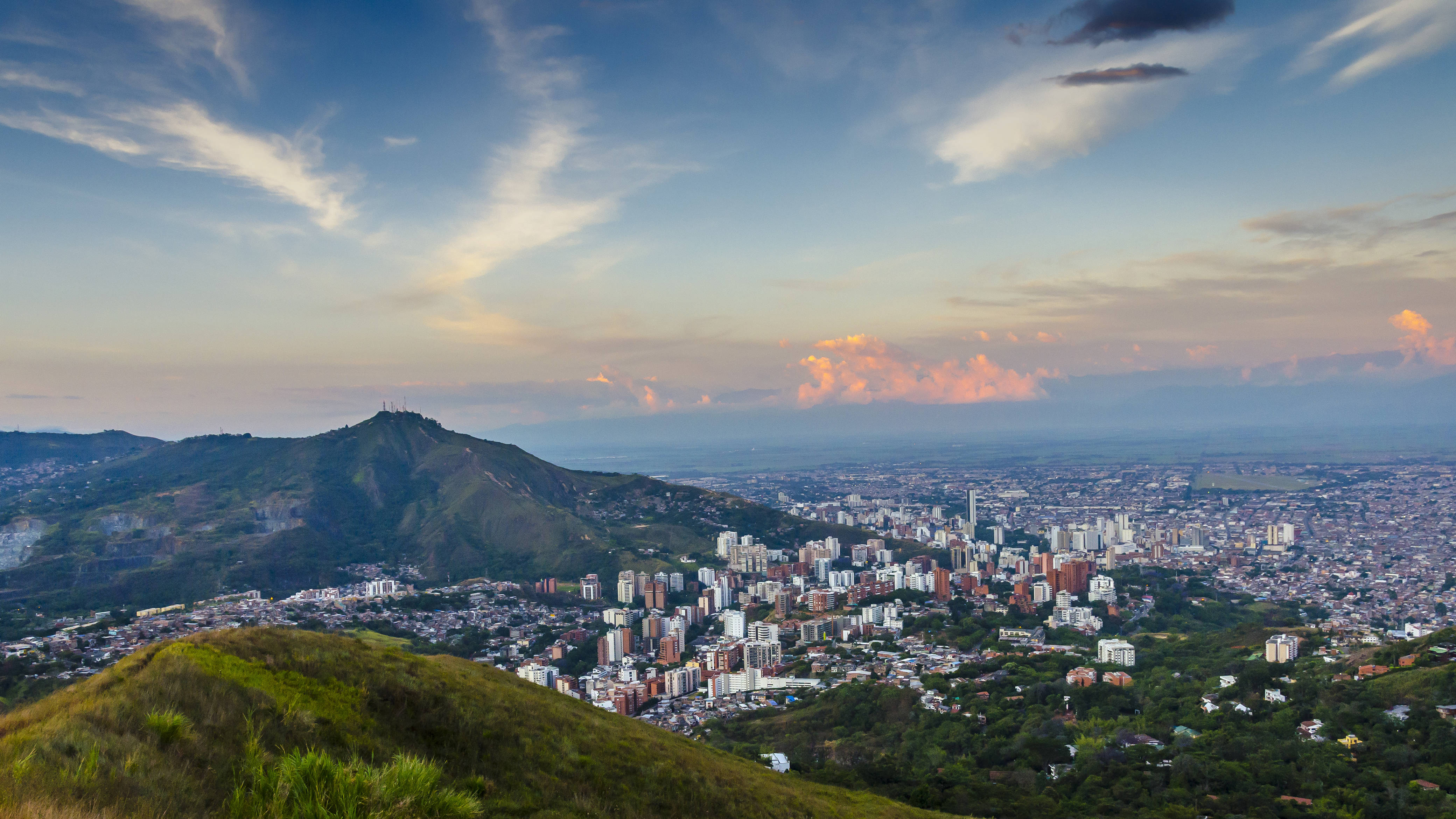 South America 桌布 壁紙 — 一座廣闊的城市坐落在山谷中，遠處有一座綠色的山在柔和的日落天空下升起。