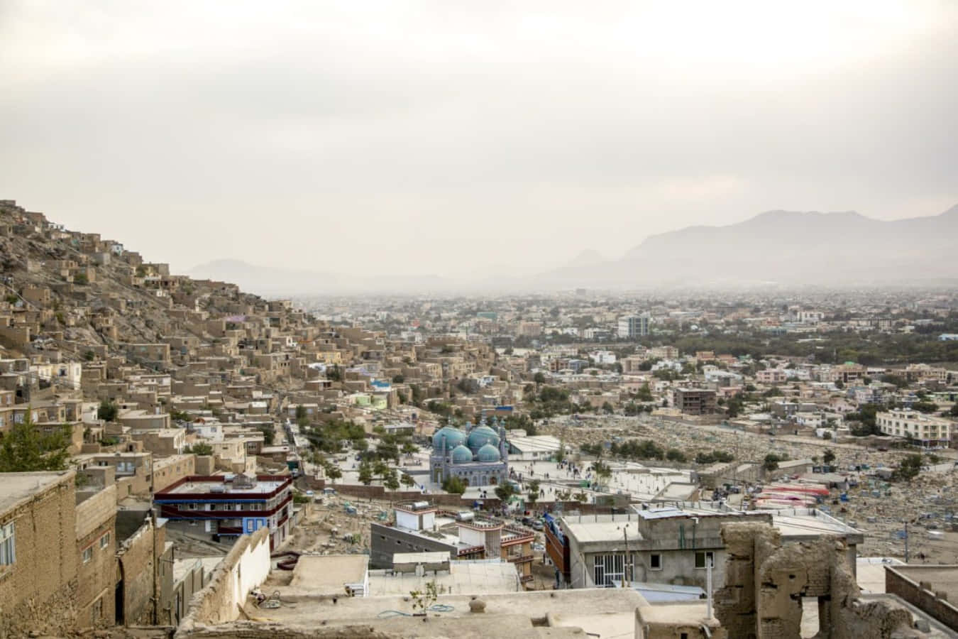 Afghanistan 桌布 壁紙 — 一座廣闊的山丘城市，擁有陶土建築和一座引人注目的藍色圓頂清真寺，天空朦朧。
