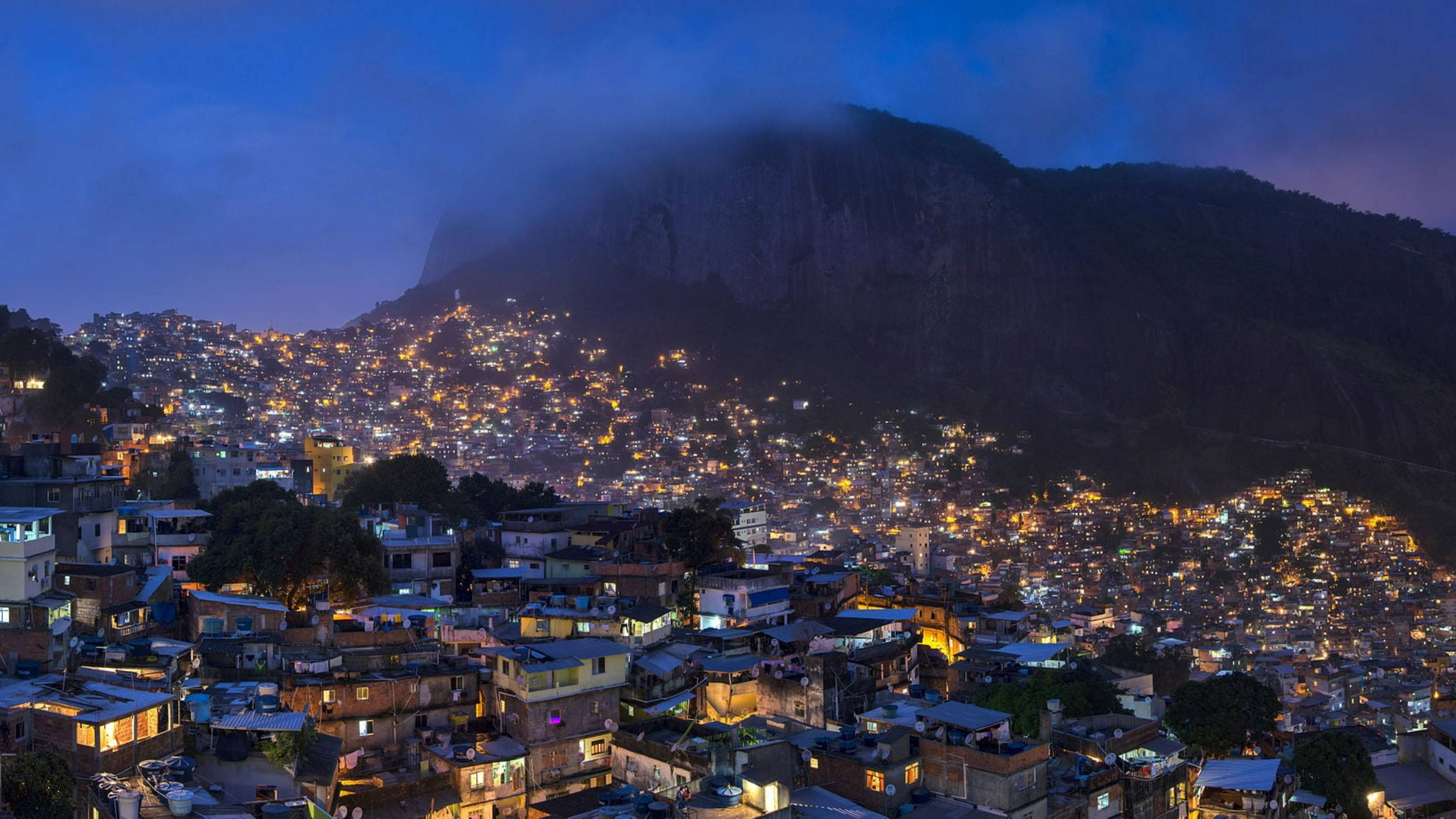 rio 桌布 壁紙 — 一座廣闊的山丘城市在暮色天空下，以溫暖燈光映照著霧氣繚繞的山巒。