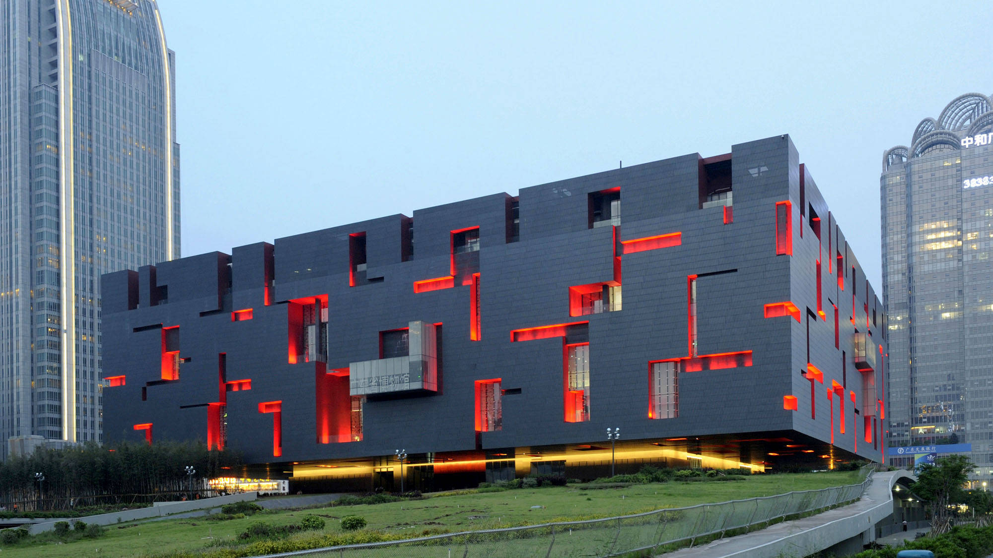 Guangzhou 桌布 壁紙 — 一座引人注目的現代建築，帶有深灰色幾何面板和發光的紅色室內燈光，在黃昏時分矗立在高樓大廈之間。